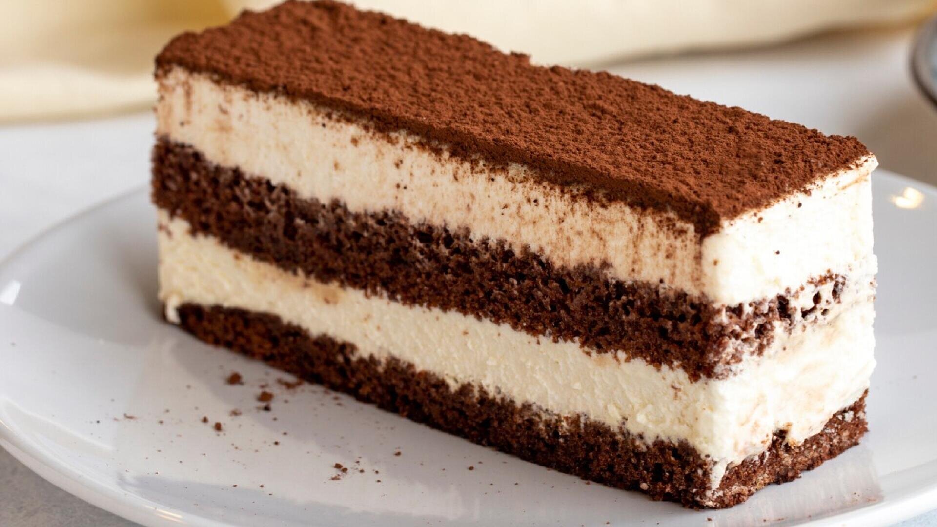 A slice of tiramisu cake with layers of creamy filling and chocolate sponge, topped with a dusting of cocoa powder, served on a white plate.