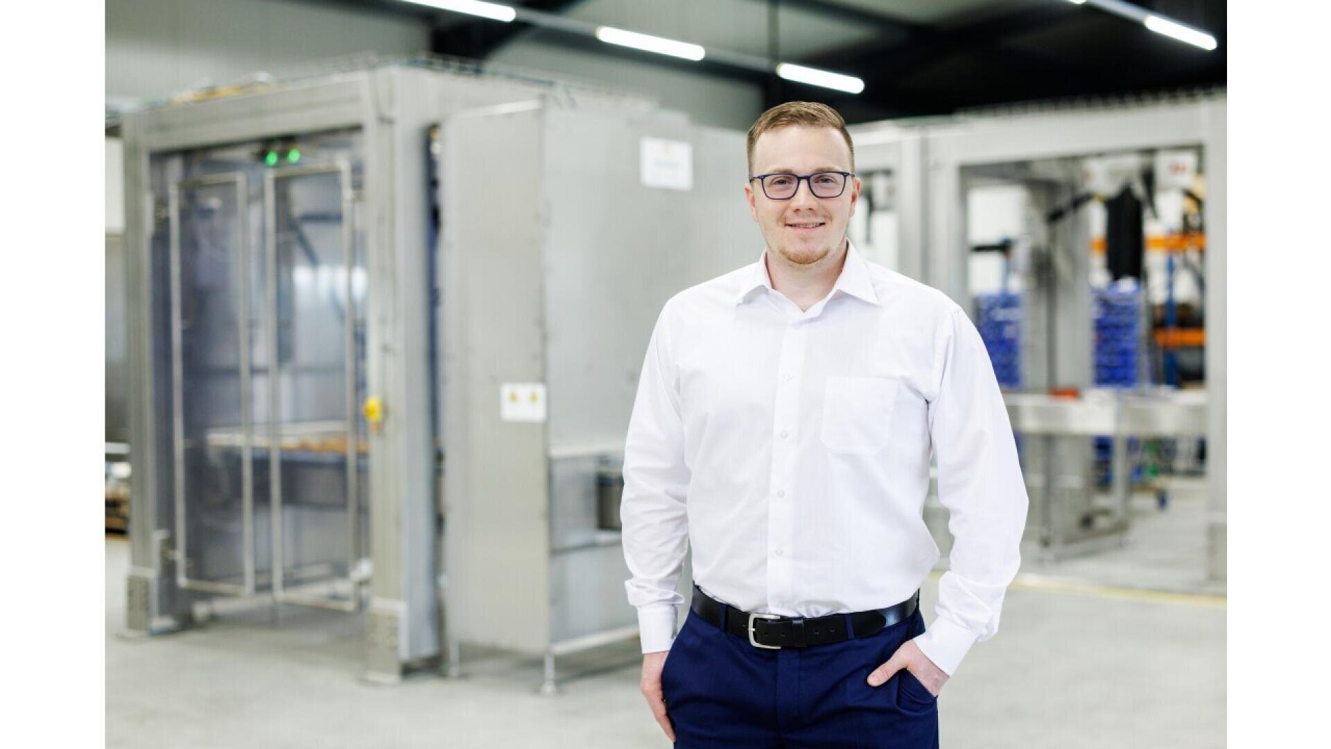 A man wearing glasses, a white shirt, and dark pants stands smiling in a modern industrial facility with large metal equipment in the background.