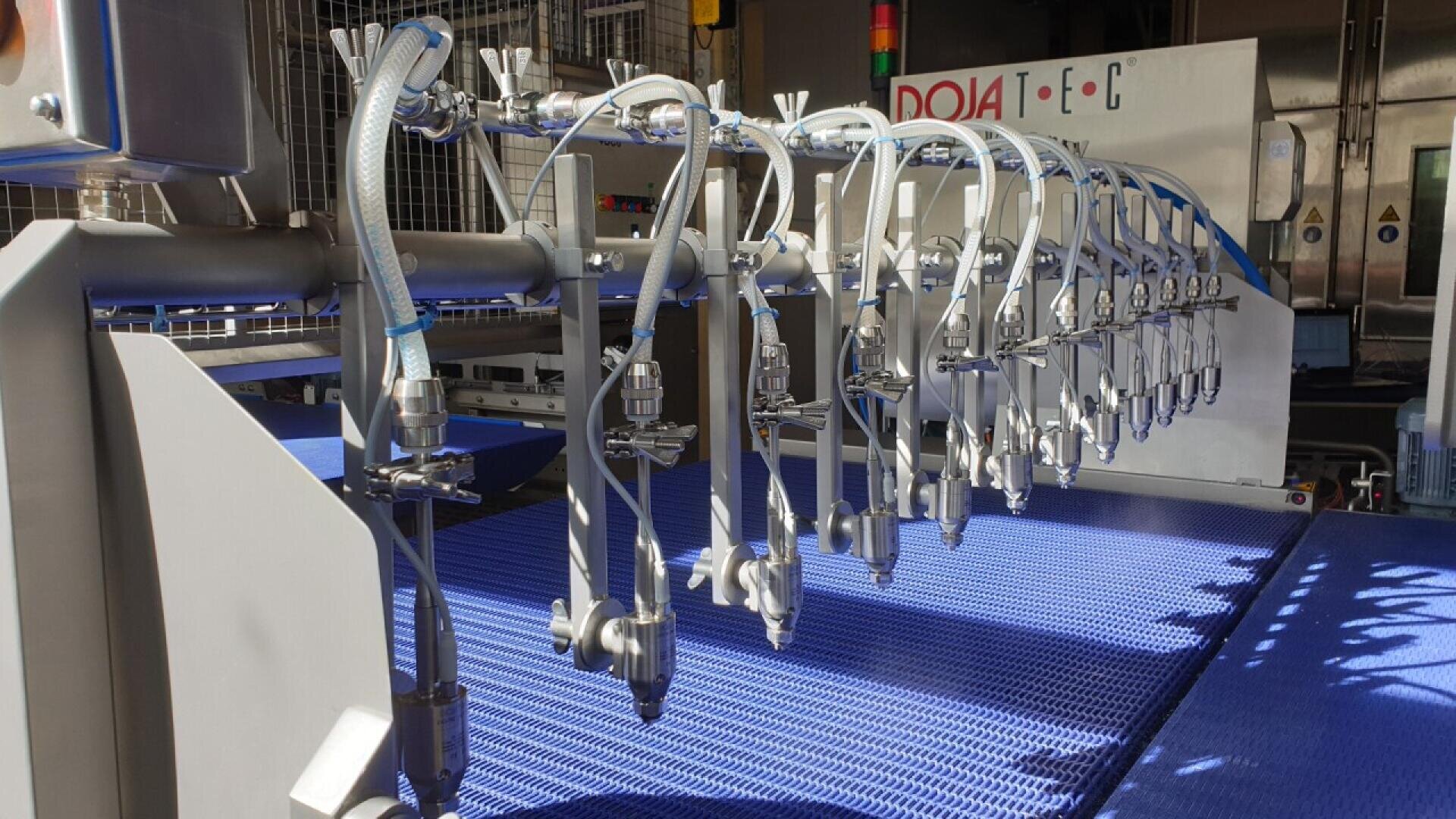 Close-up of an industrial food processing machine with multiple metal nozzles above a blue conveyor belt, likely used for automated spraying or coating in a factory setting.