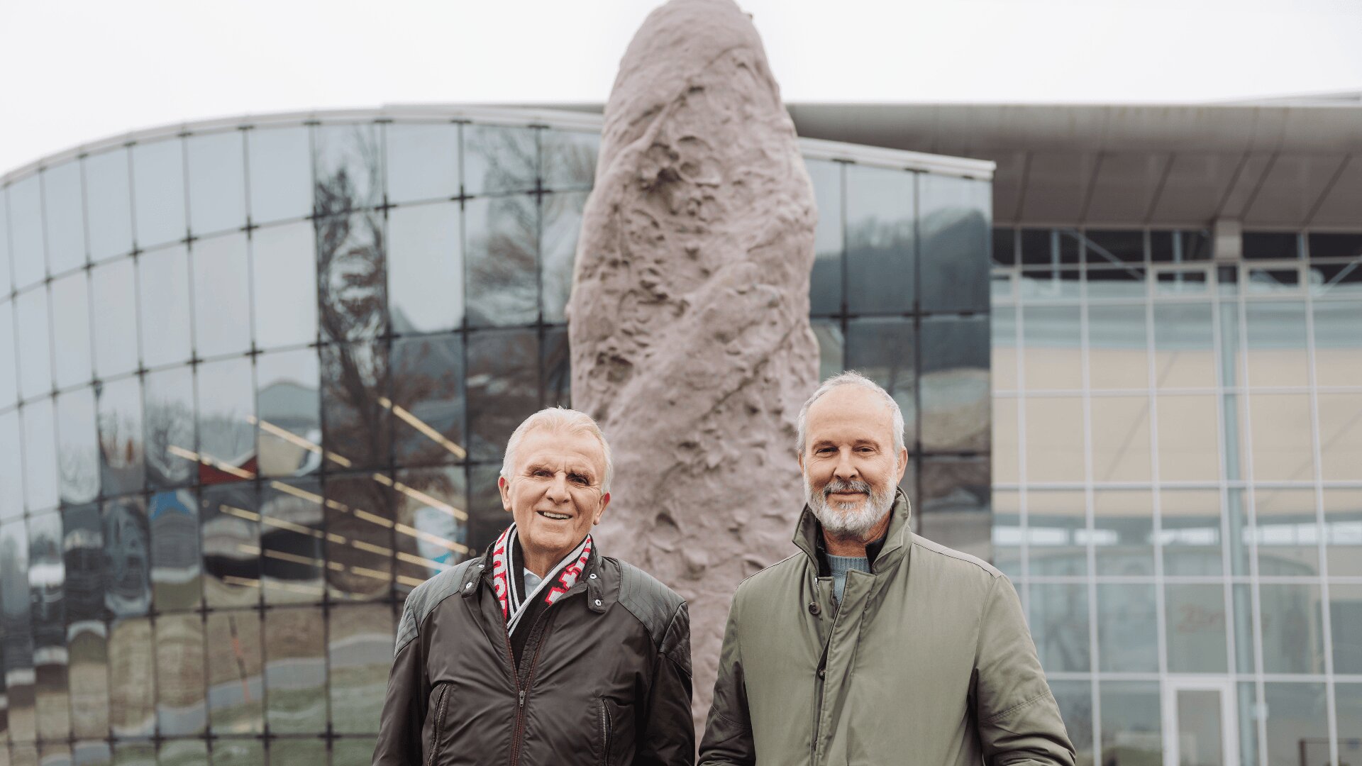 Zwei ältere Männer mit grauen Haaren und Bärten stehen lächelnd vor einem modernen Glasgebäude mit einer großen abstrakten Steinskulptur im Hintergrund. Beide tragen grüne Jacken und Schals.