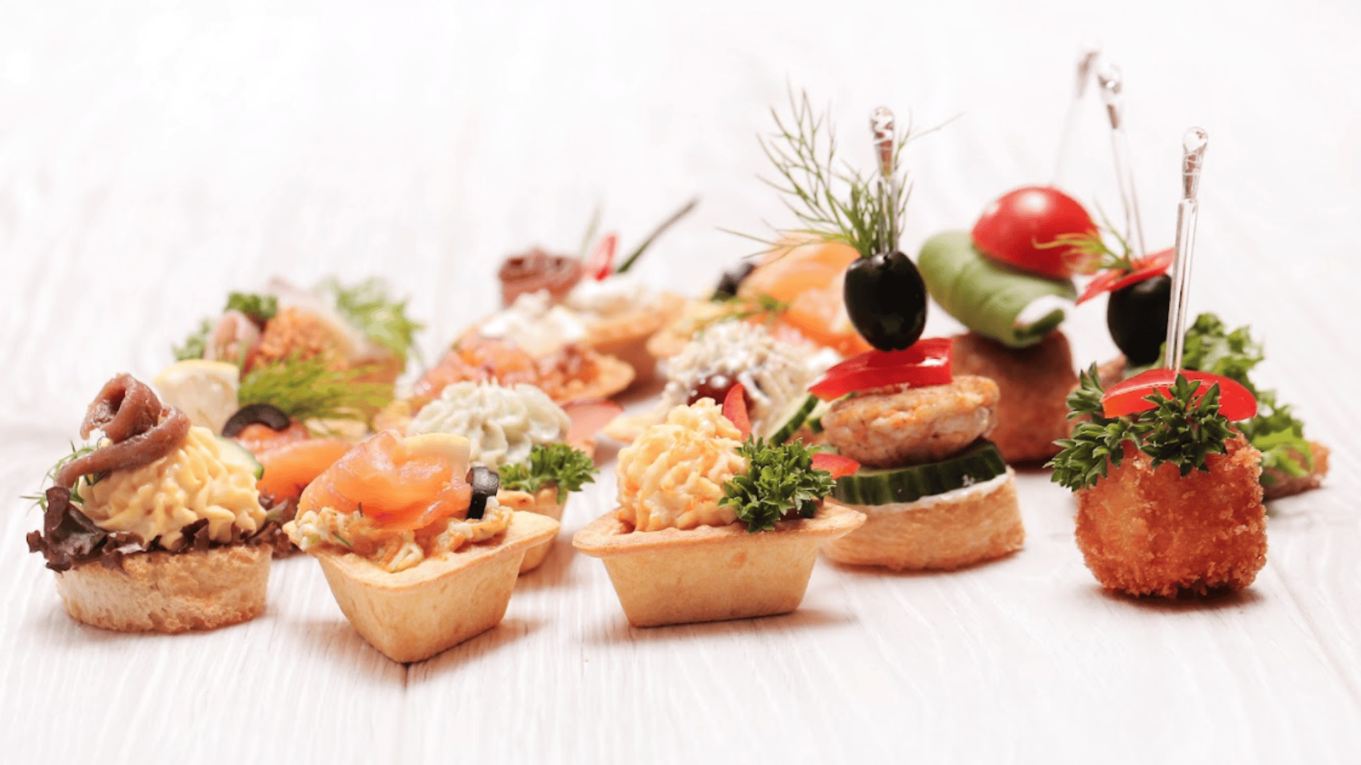 Eine Vielzahl von bunten, mundgerechten Canapés mit Zutaten wie Räucherlachs, Kräutern, Käse, Gemüse und Oliven werden auf einer hellen Holzfläche angerichtet und mit frischer Petersilie und Cocktailspießen garniert.