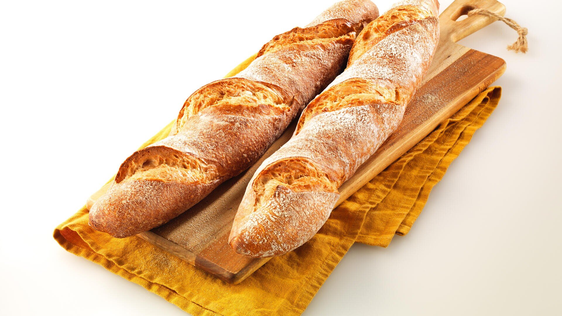 Zwei rustikale Baguettes mit knuspriger, goldbrauner Oberseite liegen auf hölzernen Schneidebrettern, die über einem gefalteten gelben Tuch auf weißem Hintergrund liegen.