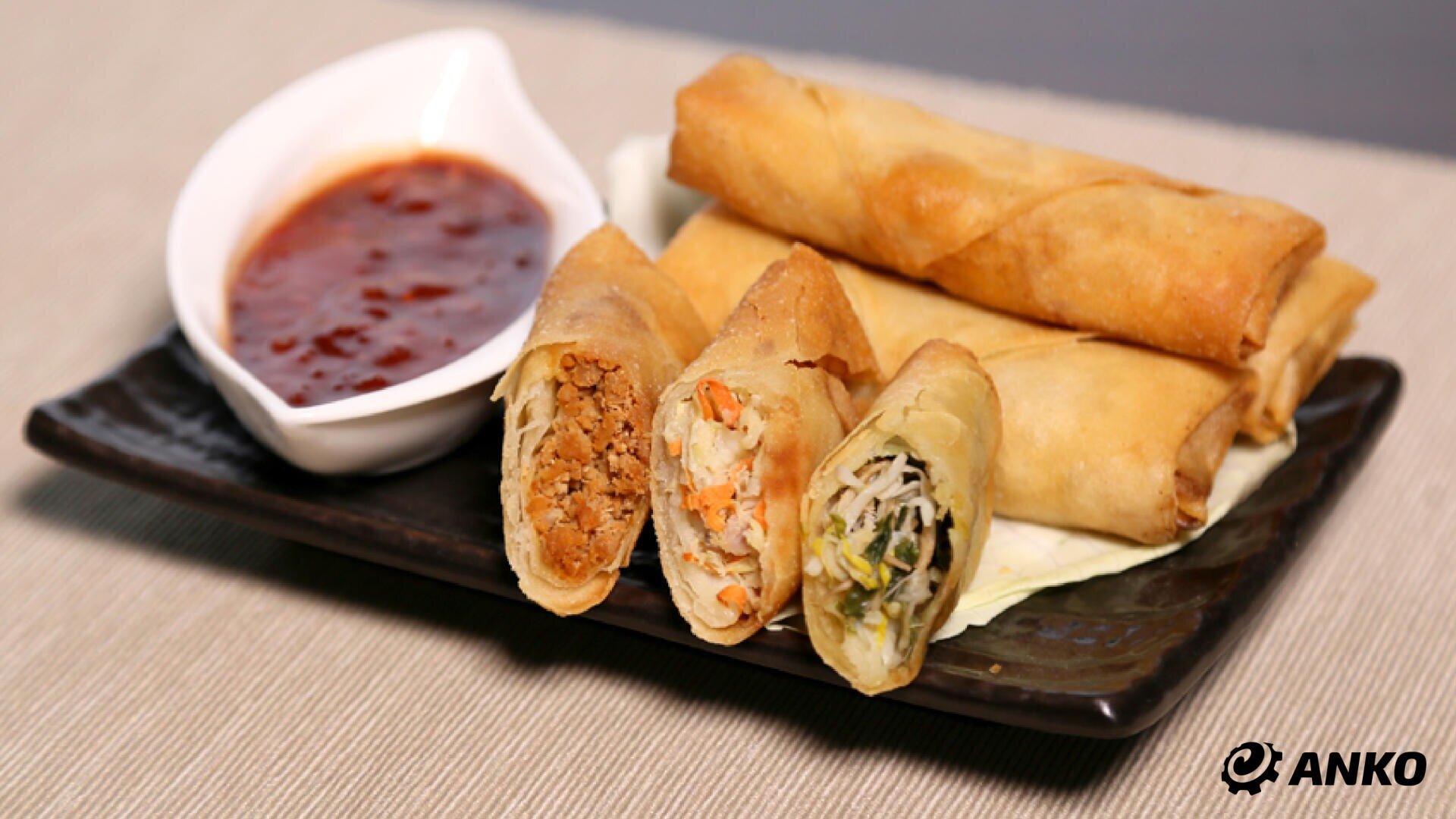 A black plate of golden fried spring rolls, some of which are cut open to reveal vegetable and meat fillings, next to a small white bowl of red dipping sauce. The ANKO logo can be seen in the corner.