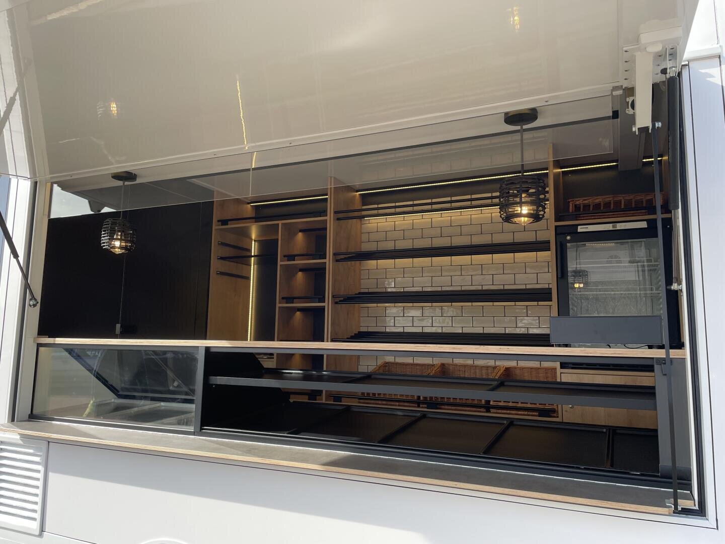 Ein Foodtruck mit geöffnetem Servicefenster, das einen leeren Innenraum mit Holzregalen, einer gefliesten Aufkantung und hängenden Pendelleuchten zeigt. Im Inneren sind keine Lebensmittel, Geräte oder Mitarbeiter zu sehen.