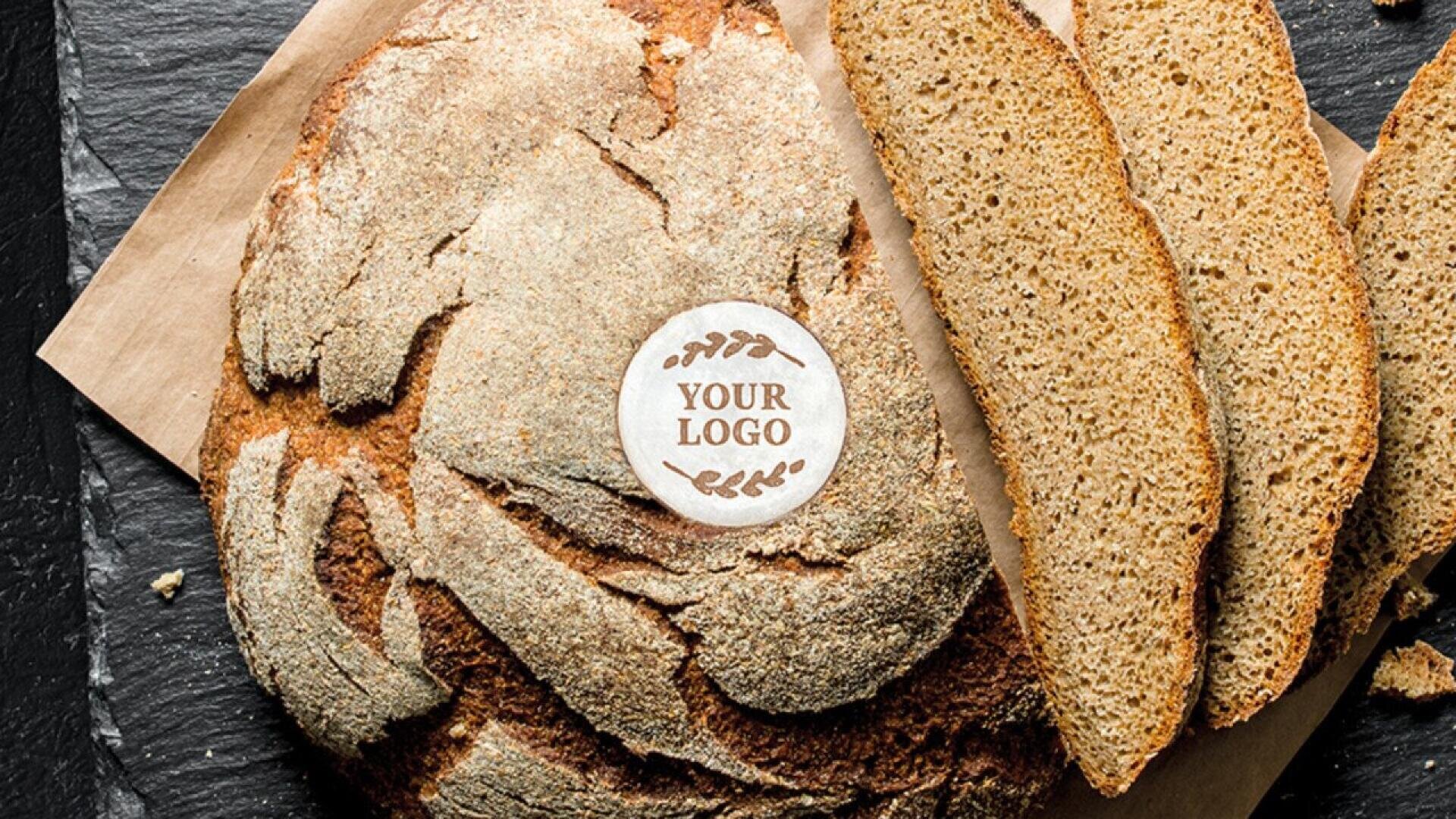 A round rustic loaf of bread with a custom logo label in the center, partially sliced, displayed on parchment paper and a dark slate surface.
