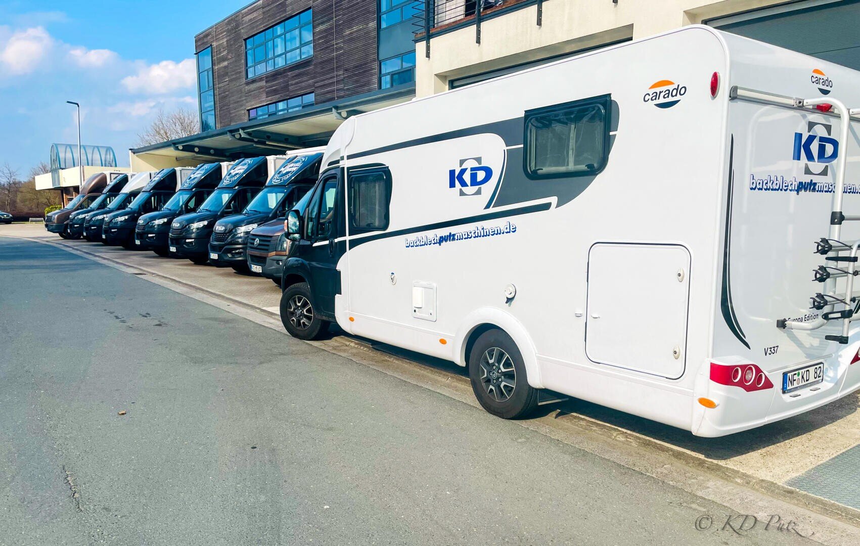 An einem sonnigen Tag parkt ein weißer Wohnmobil der Marke KD neben einer Reihe mehrerer schwarzer Wohnmobile der Marke KD vor einem modernen Gebäude.