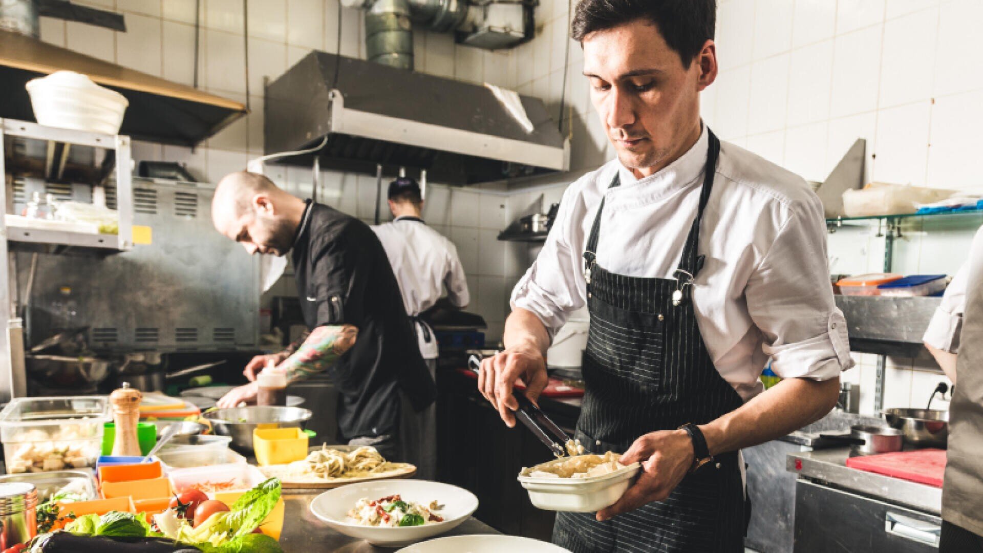 Two chefs in a commercial kitchen, both wearing aprons. One chef is plating food into a takeout container, while the other works in the background near a stove. Various ingredients and dishes are on the counter.