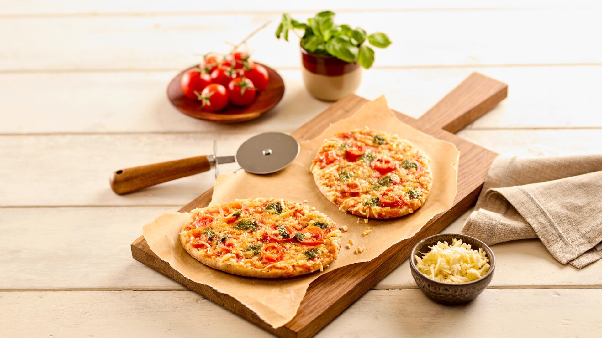 Zwei Fladenbrotpizzen mit Tomaten, Käse und Kräutern auf Pergamentpapier auf einem Holzbrett. In der Nähe stehen ein Pizzaschneider, eine Schüssel mit geriebenem Käse, ein Topf mit Basilikum und ein Teller mit Kirschtomaten.