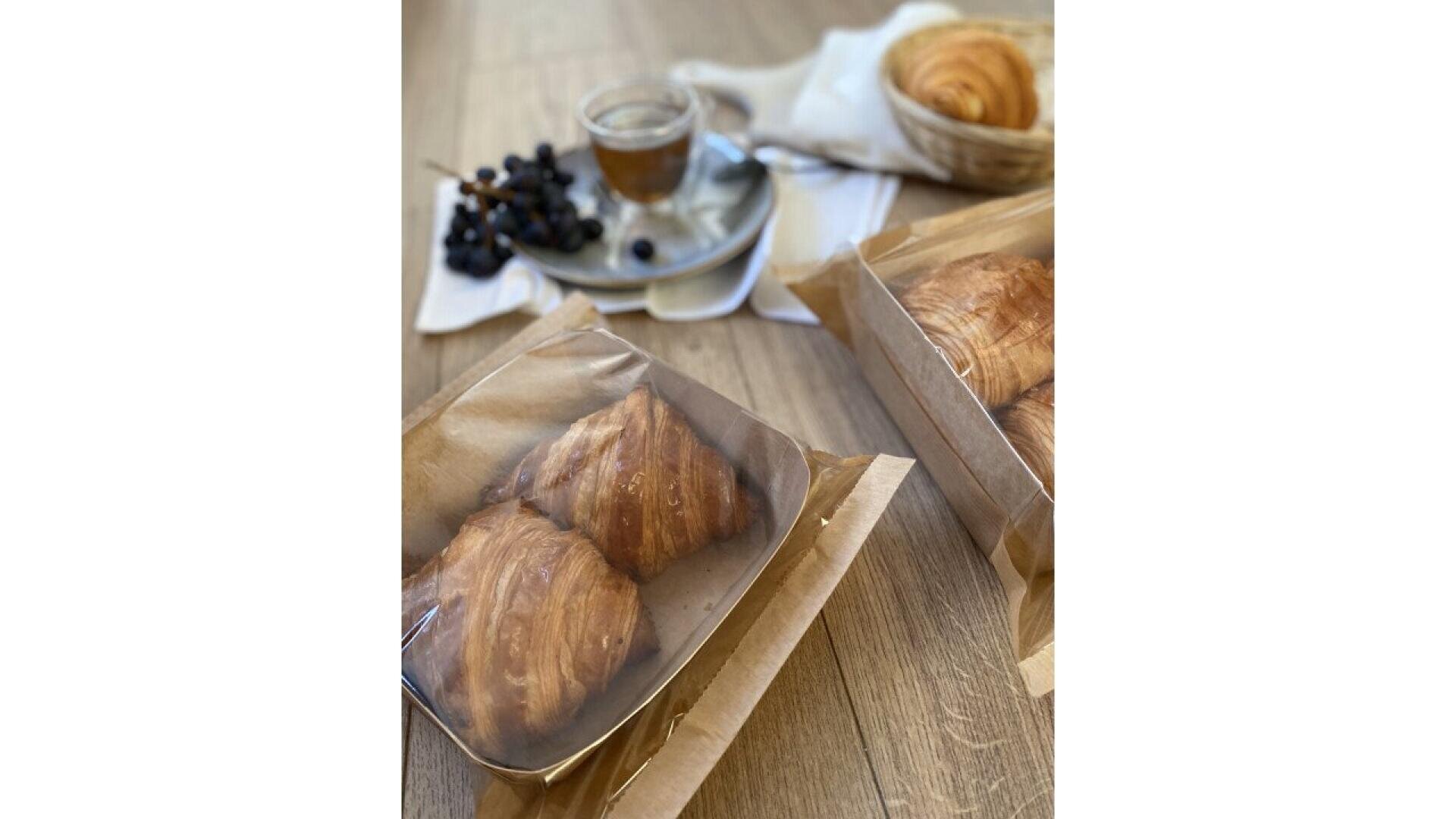 Zwei Schachteln mit Croissants auf einem Holztisch, mit Weintrauben, einer Tasse Kaffee und einem Korb mit weiteren Croissants im Hintergrund. Die Szene ist hell und einladend.