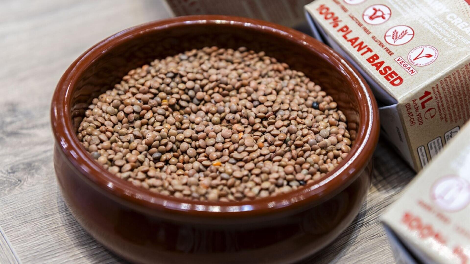 A brown ceramic bowl filled with dried lentils sits on a wooden surface, with plant-based food packaging labeled 100% PLANT BASED in the background.