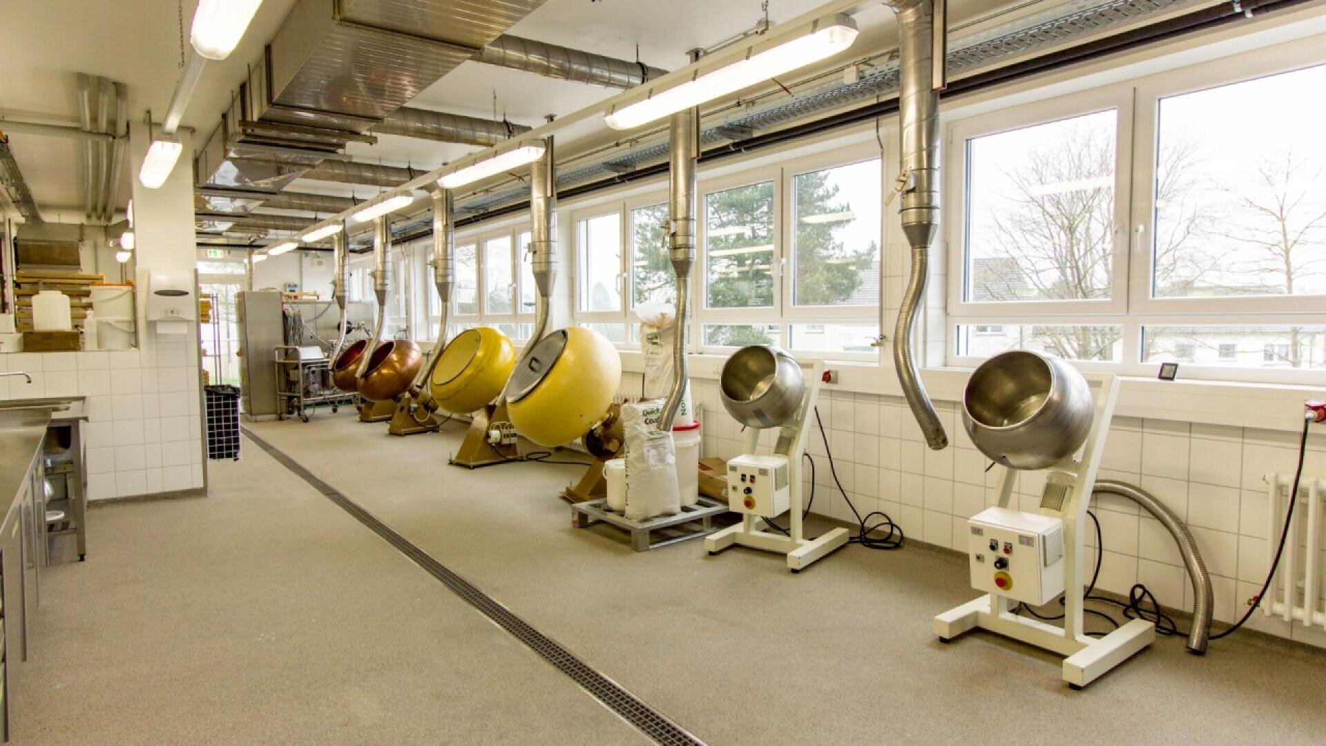 A clean, modern industrial kitchen with large windows, multiple metal mixing or coating machines lined up, and overhead ductwork. The room is well-lit, spacious, and organized.