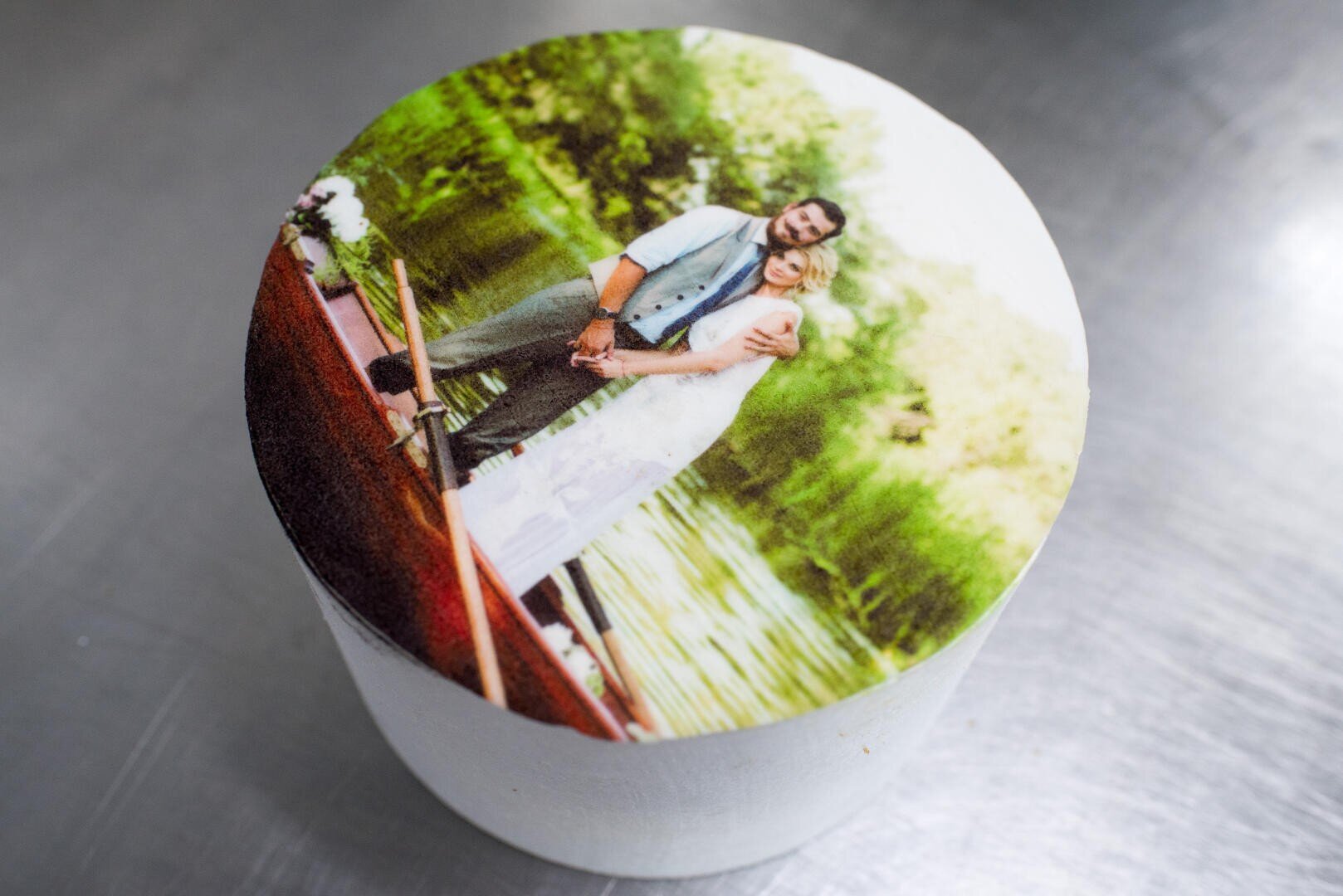 A round cake with a printed edible image of a couple in wedding attire standing on a boat, surrounded by greenery and water, placed on a metallic surface.
