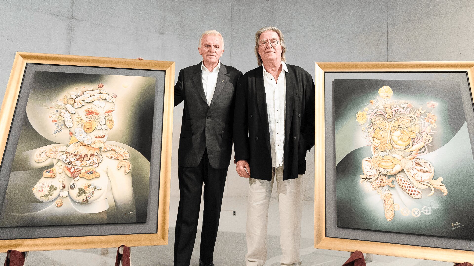 Zwei Männer in Anzügen stehen in einem Innenraum nebeneinander, jeder neben einem großen gerahmten abstrakten Gemälde, das bunte, surreale humanoide Figuren zeigt. Der Hintergrund ist eine schlichte, hellgraue Wand.