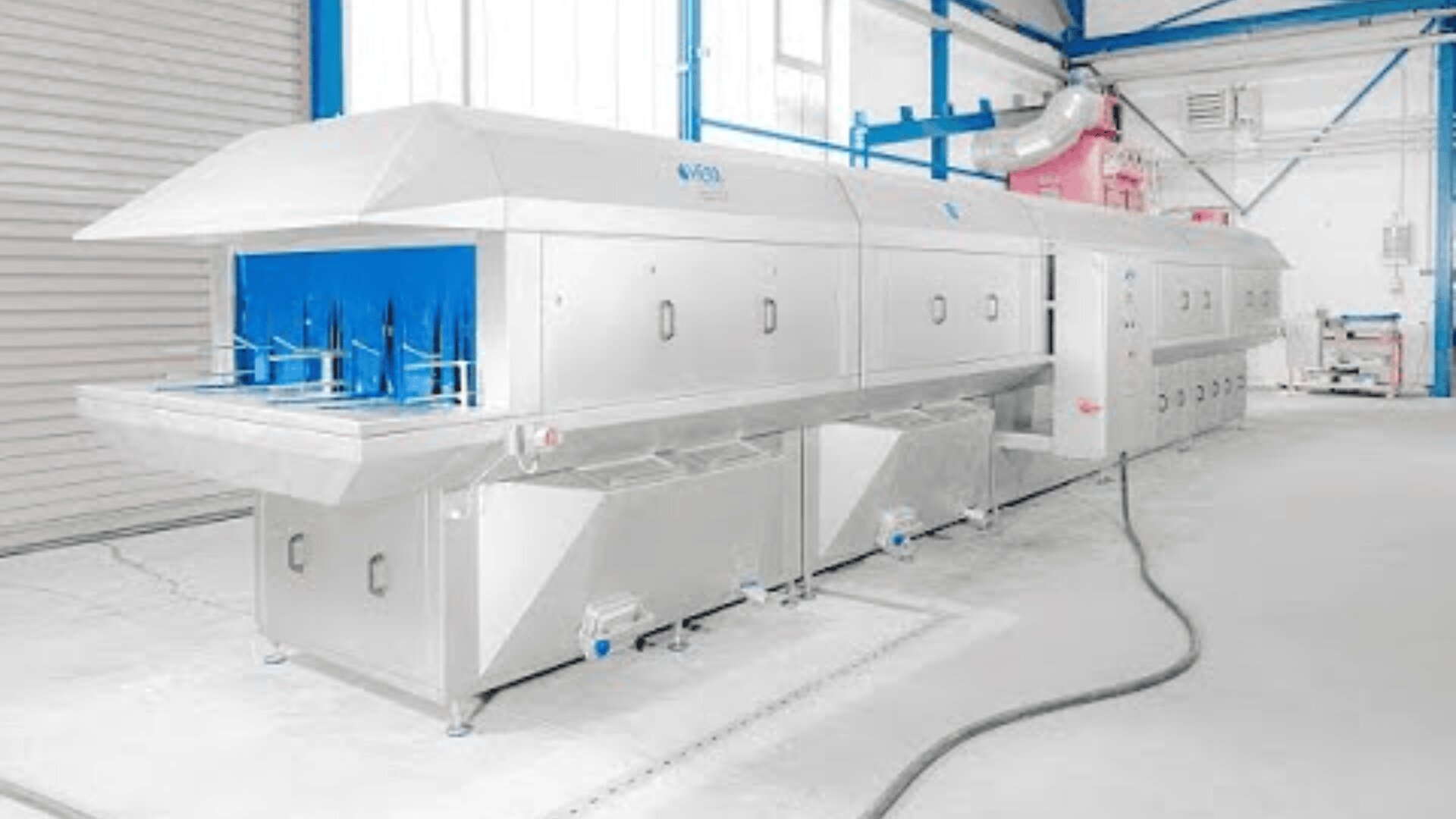 Large industrial machine with a stainless steel exterior and blue accents, located inside a factory setting; pipes, vents, and cables are visible around the equipment.