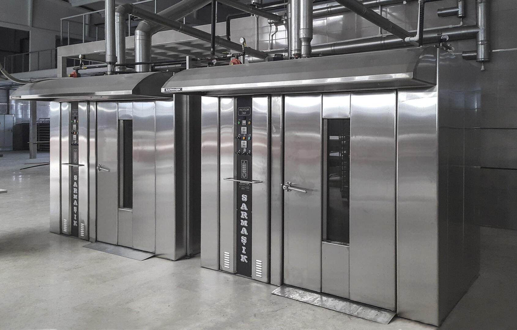 Two large, industrial stainless steel rotary ovens with glass windows, control panels, and exhaust pipes are positioned side by side in a commercial bakery or food production facility.