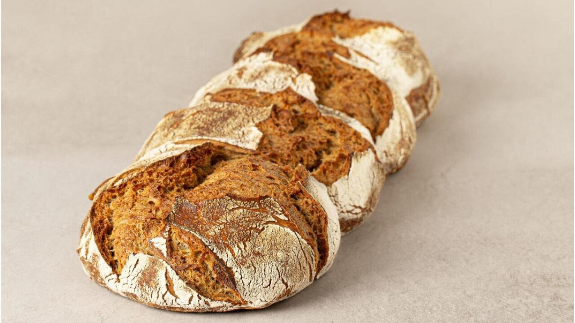Four round loaves of rustic, crusty bread with cracked, flour-dusted tops are lined up diagonally on a light beige surface.