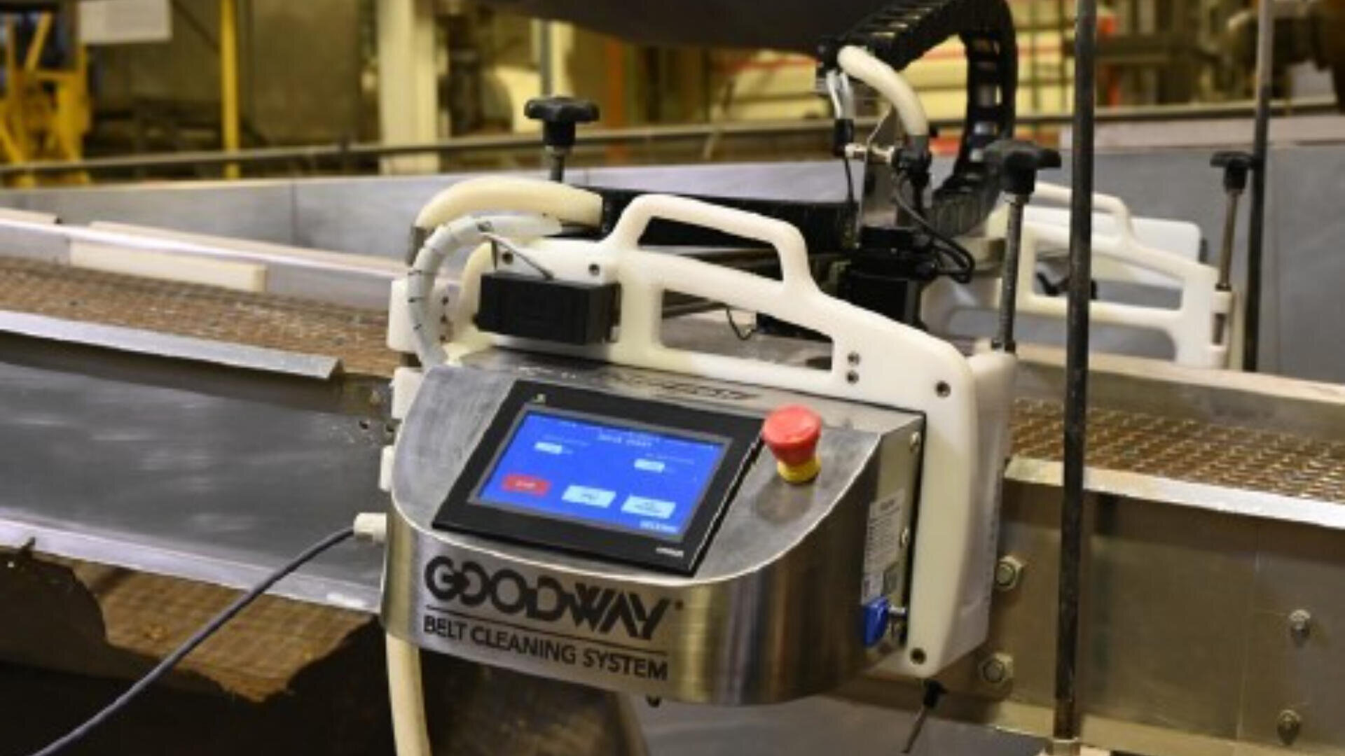Ein Goodway-Bandreinigungssystem mit einer Touchscreen-Oberfläche und einer Not-Aus-Taste ist an einem industriellen Förderband in einer Fabrik angebracht.