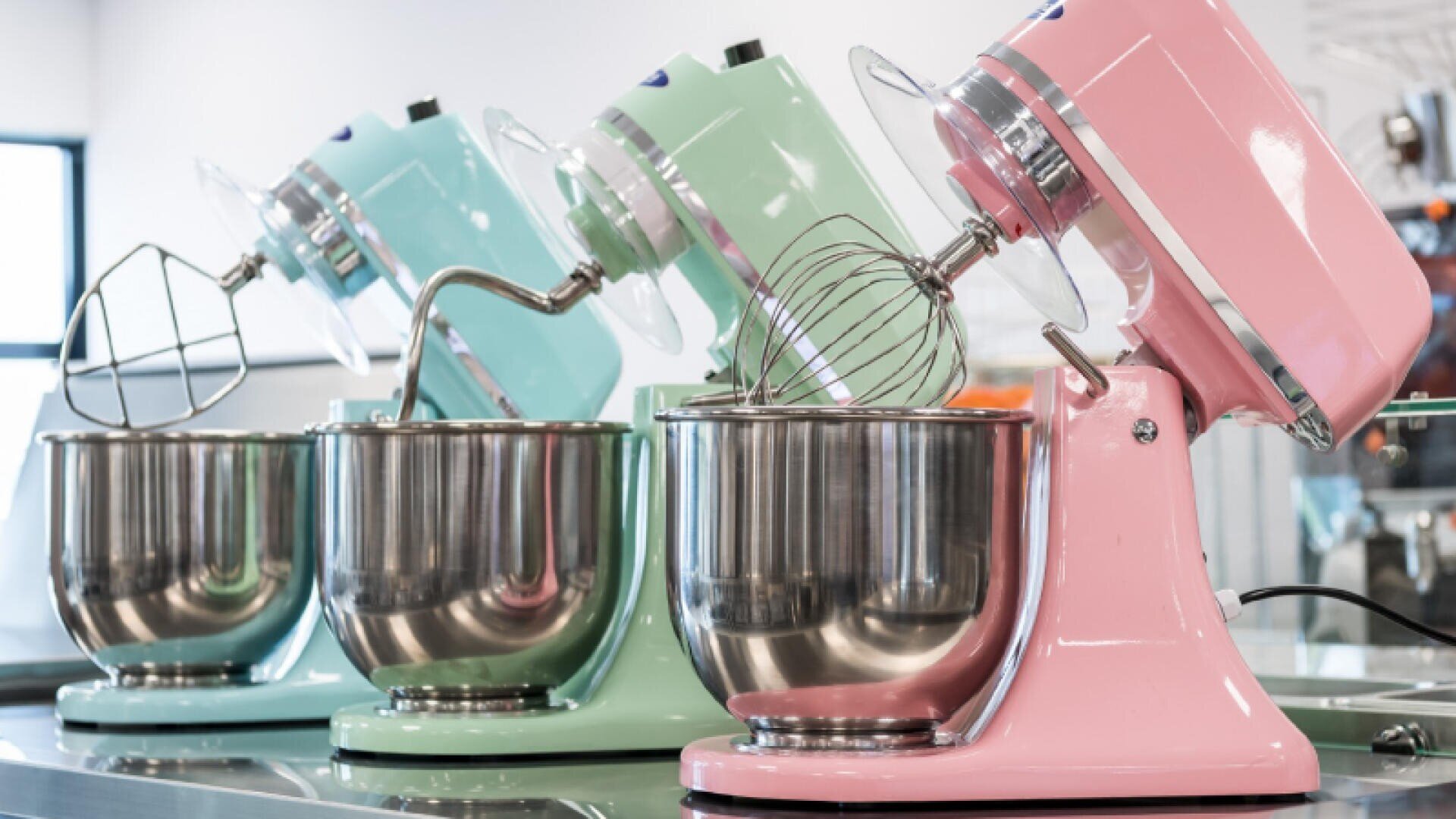 Three pastel-colored stand mixers—blue, mint green, and pink—sit in a row on a countertop, each with a metal mixing bowl and different attachments. The background shows a bright, modern kitchen setting.