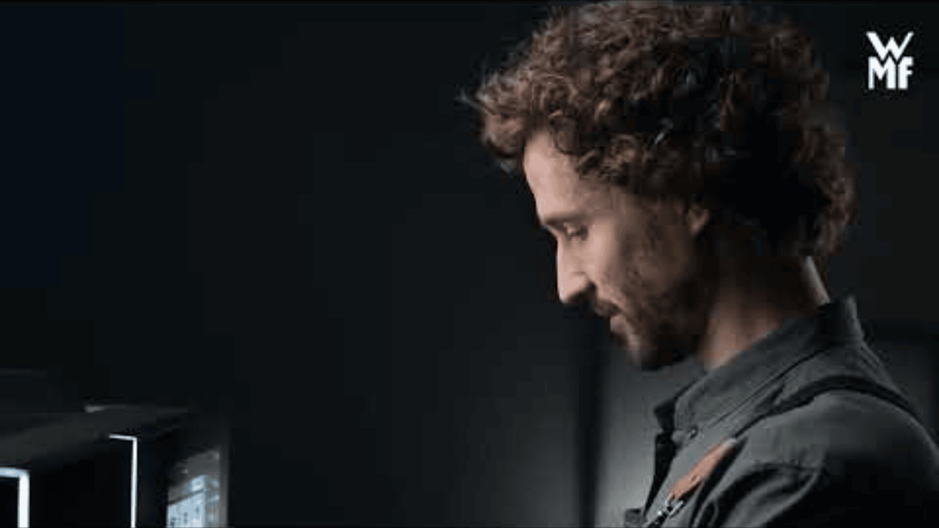 A man with curly hair and a beard looks down and smiles next to a coffee machine in a dimly lit setting. The WMF logo appears in the top right corner.
