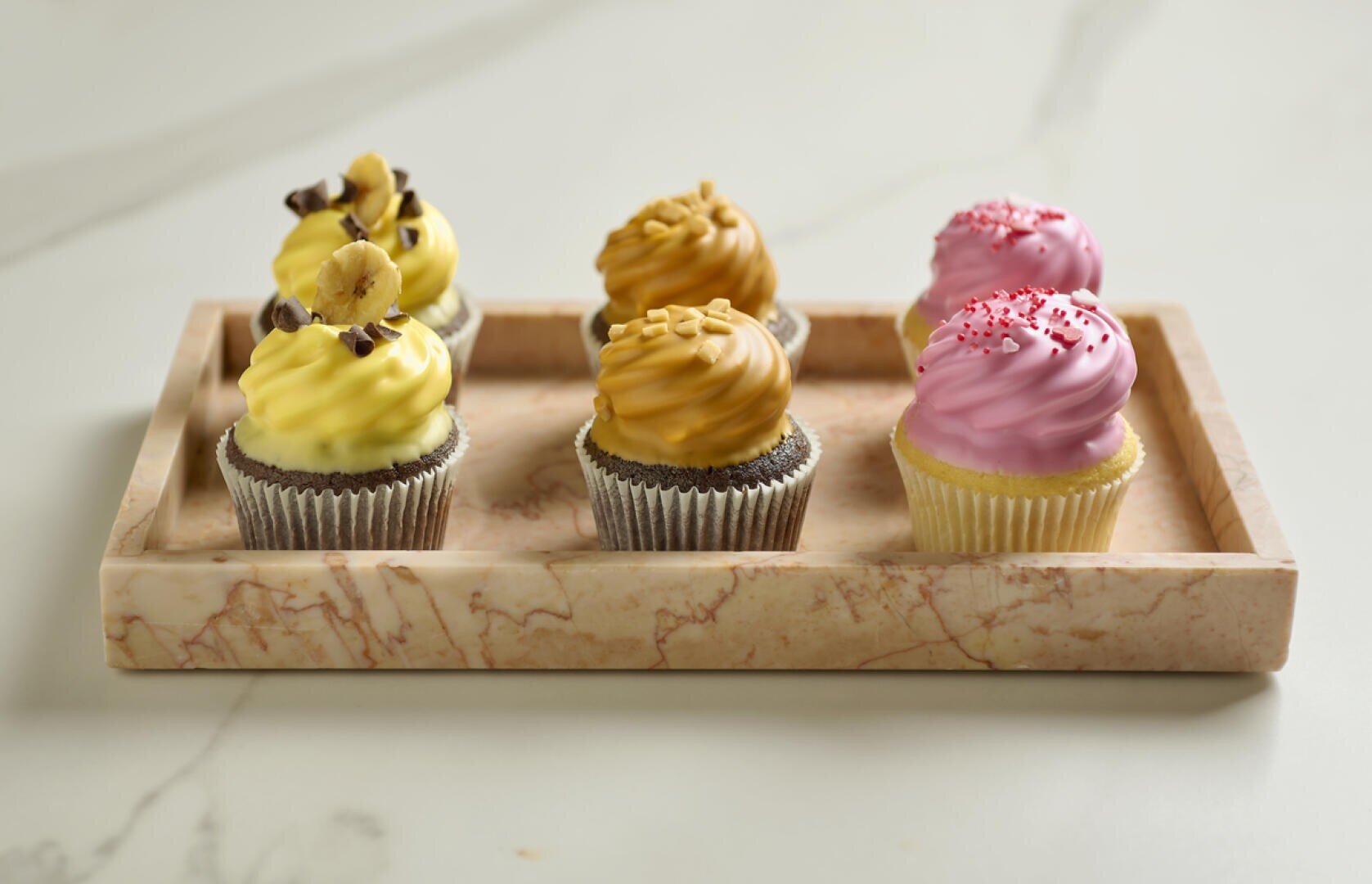 A marble tray holds six cupcakes: two with yellow frosting and chocolate toppings, two with caramel-colored frosting, and two with pink frosting and red sprinkles, all in metallic cupcake liners.