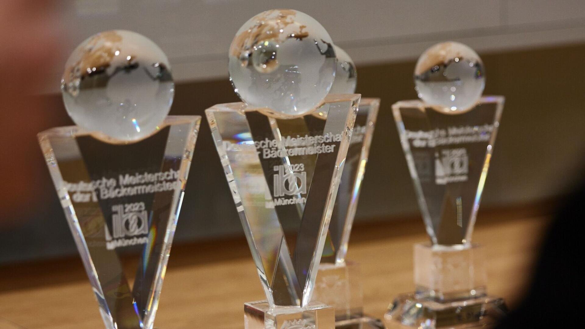 Three crystal trophies with globe-shaped tops and engraved text are displayed on a wooden surface, representing awards for the 2023 Deutsche Meisterschaft der Bäckermeister in München.