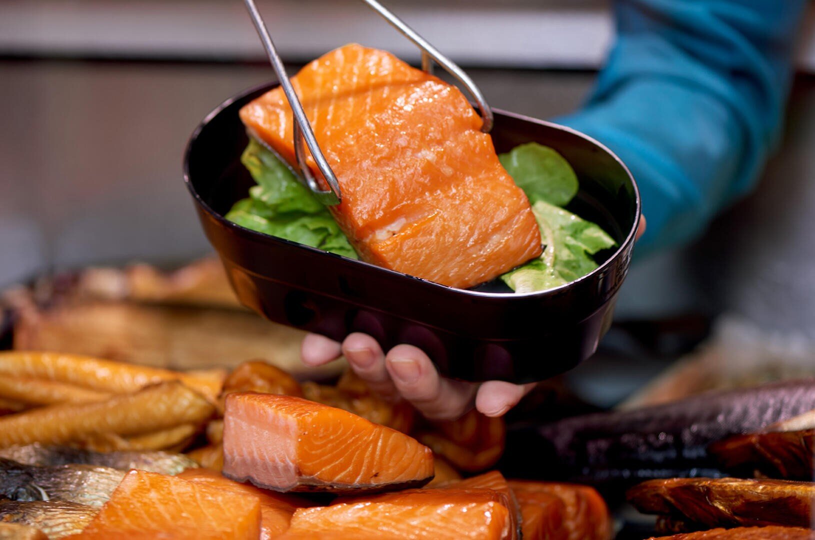 Eine Person benutzt eine Zange, um ein Stück gekochten Lachs auf einem Bett aus Blattgemüse in einer schwarzen Schale zu platzieren, wobei weitere Lachsstücke auf dem Tisch darunter zu sehen sind.