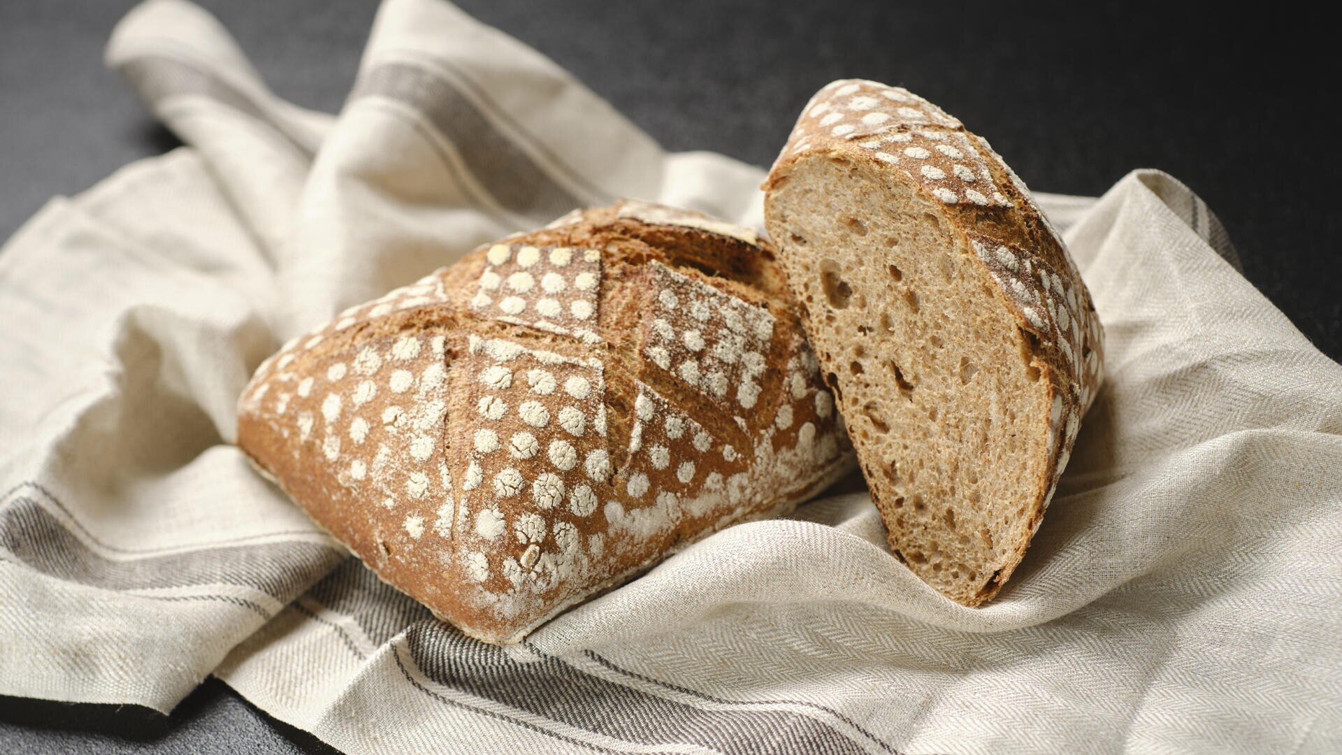 A rustic loaf of bread with a patterned flour crust lies on a beige and gray striped cloth, one slice cut off to reveal the soft, airy crumb inside.