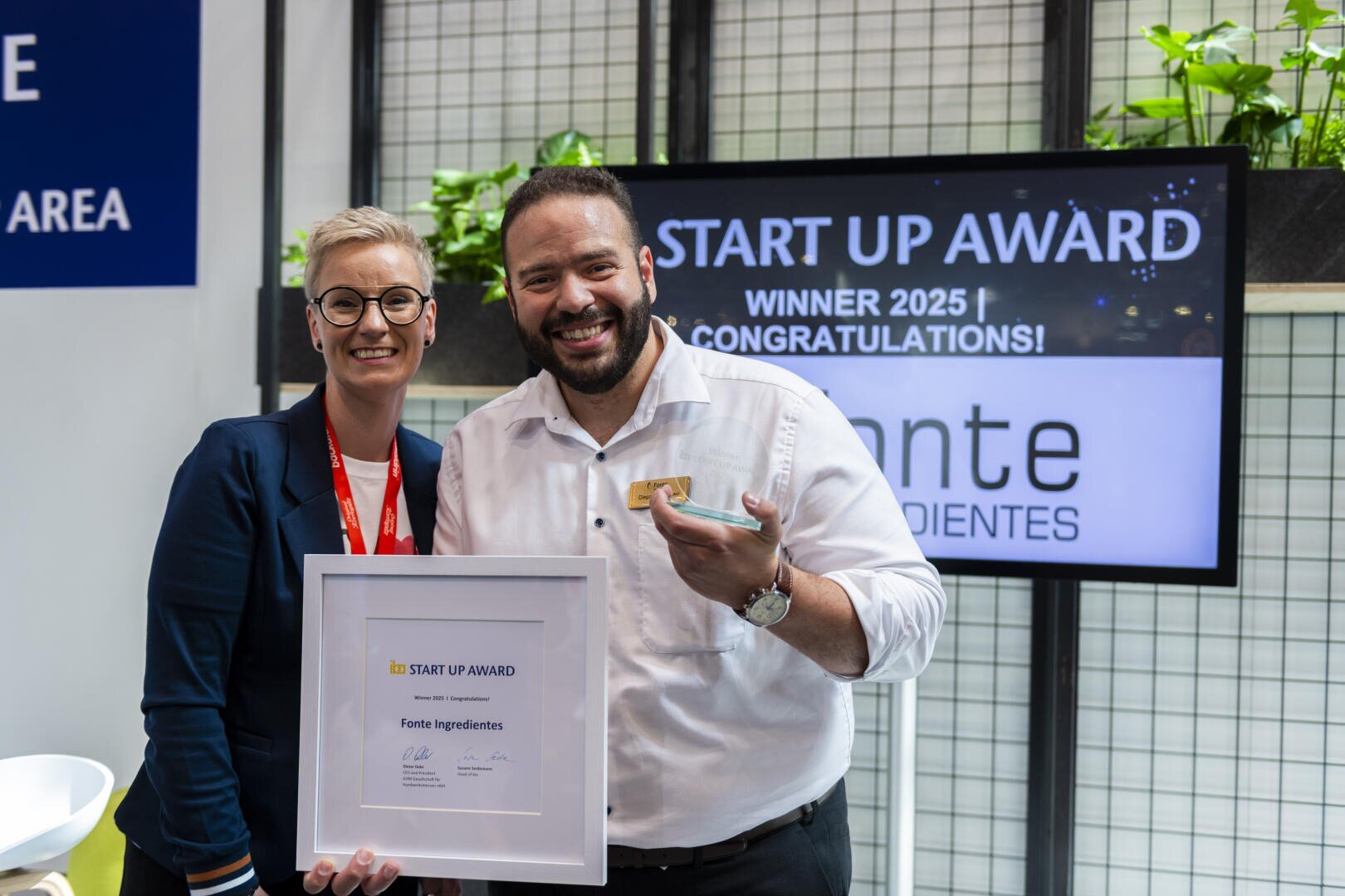 Two smiling people hold a framed certificate in front of a screen displaying START UP AWARD WINNER 2025! CONGRATULATIONS! at an event. The man is also holding a small plate. Plants decorate the background.