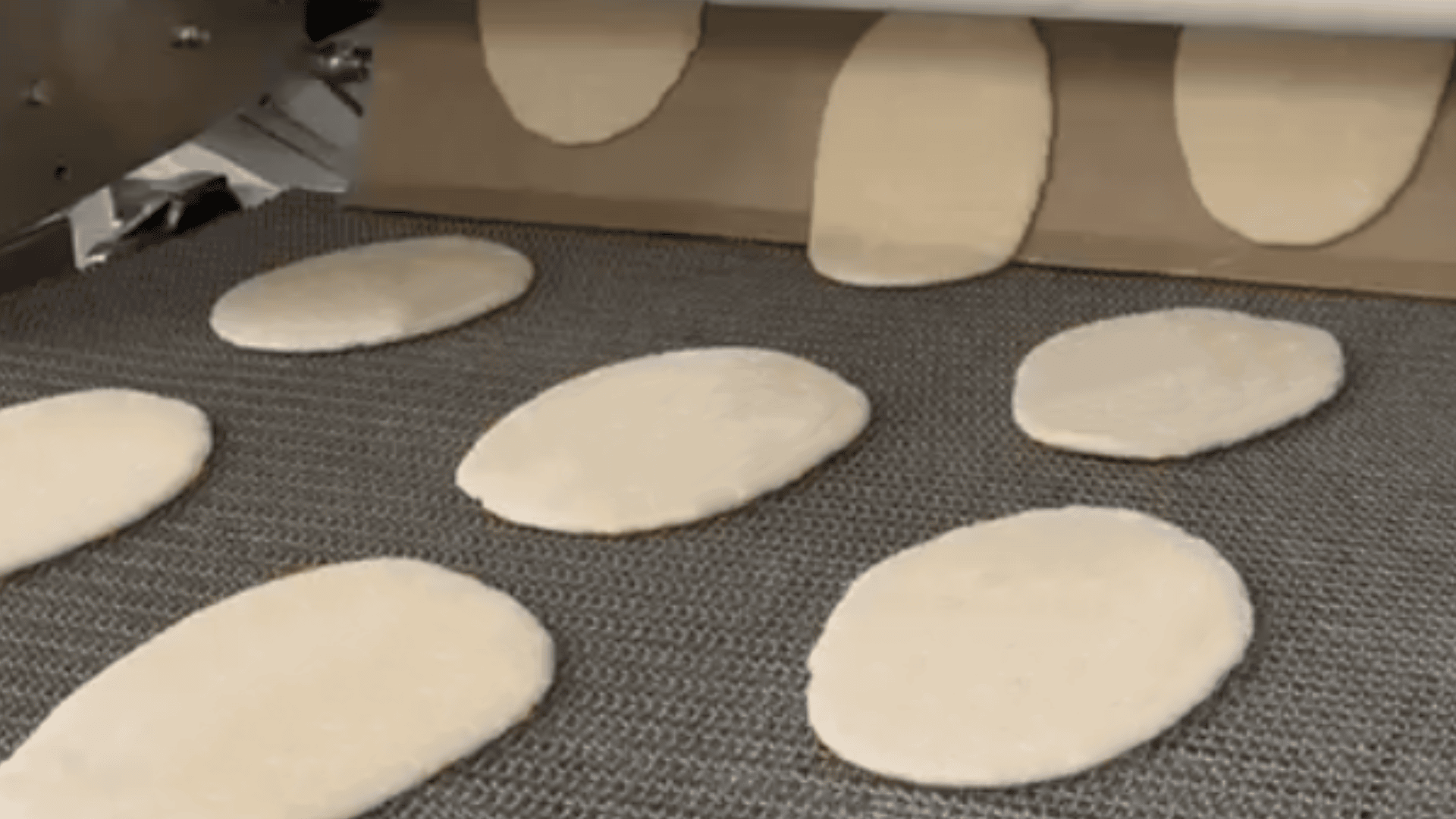 Oval pieces of dough are being dispensed onto a conveyor belt by a machine, likely as part of an automated process in a bakery or food production facility.
