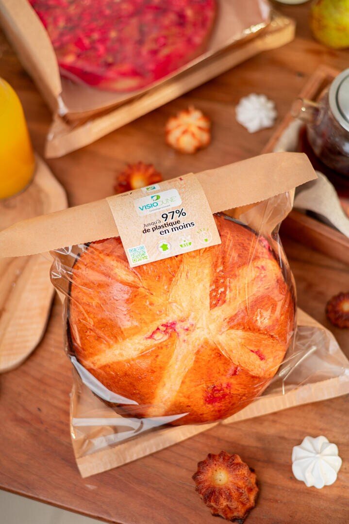 A round loaf of bread in a clear plastic bag with a paper label sits on a wooden table, surrounded by baked goods and jars. The label shows organic certification and 97%, possibly indicating purity or content.