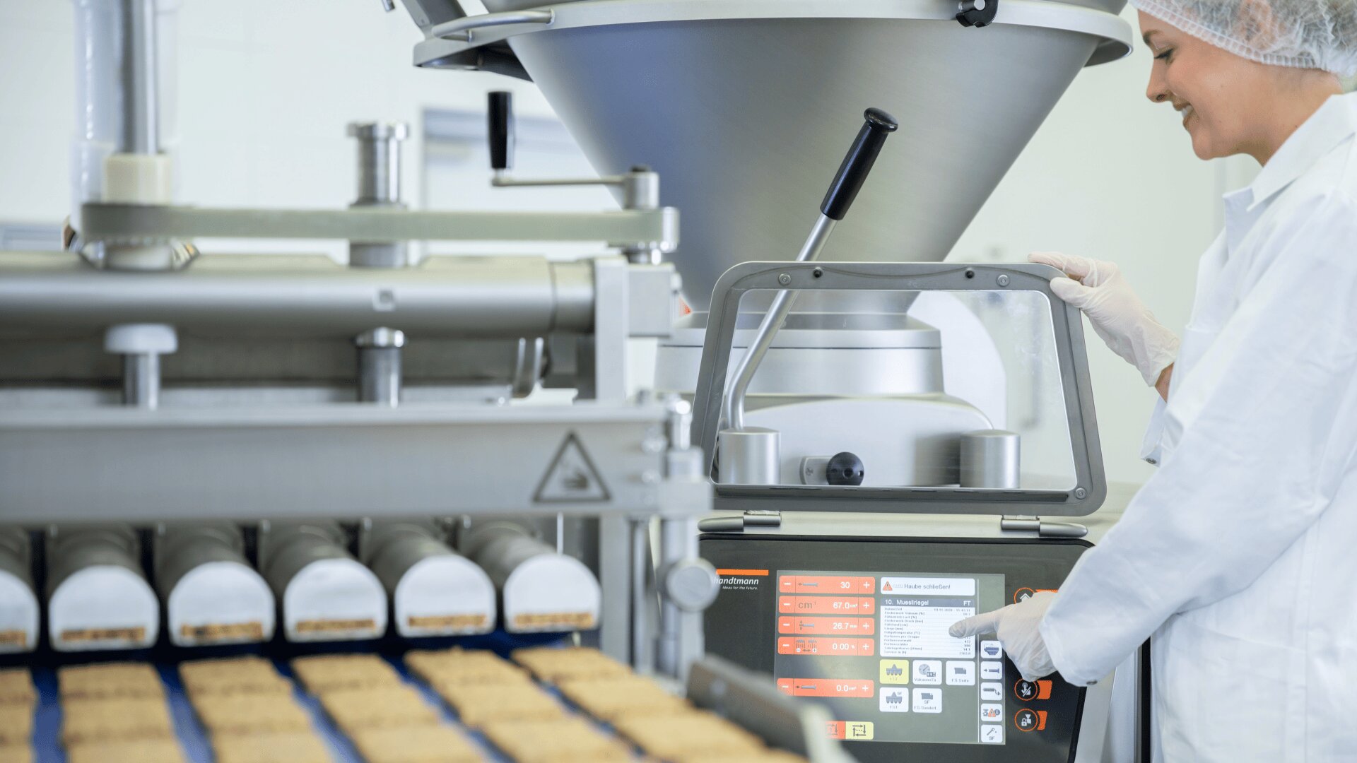 Ein Arbeiter in Laborkittel und Haarnetz bedient ein Touchscreen-Bedienfeld neben einer Lebensmittelverarbeitungsmaschine, die in einer sauberen Industrieumgebung rechteckige Riegel auf einem Förderband produziert.