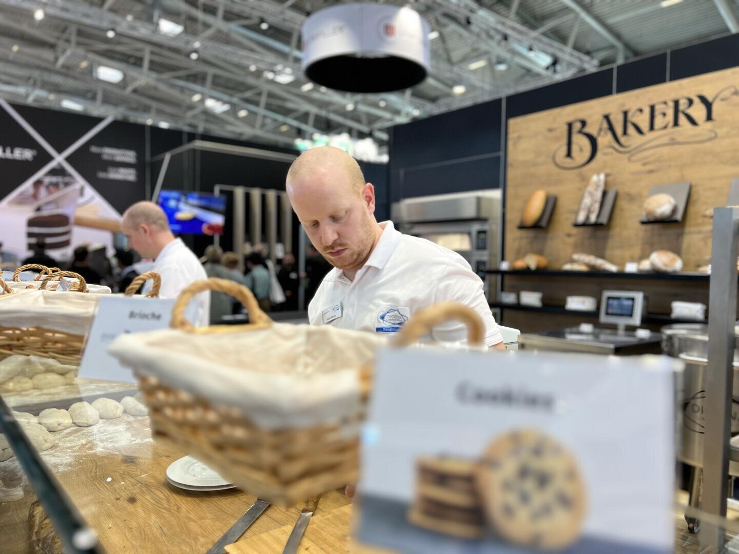 A man in a white shirt works behind a bakery counter with baskets of dough and pastries. A sign labeled Cookies is in the foreground, and shelves with bread are in the background.