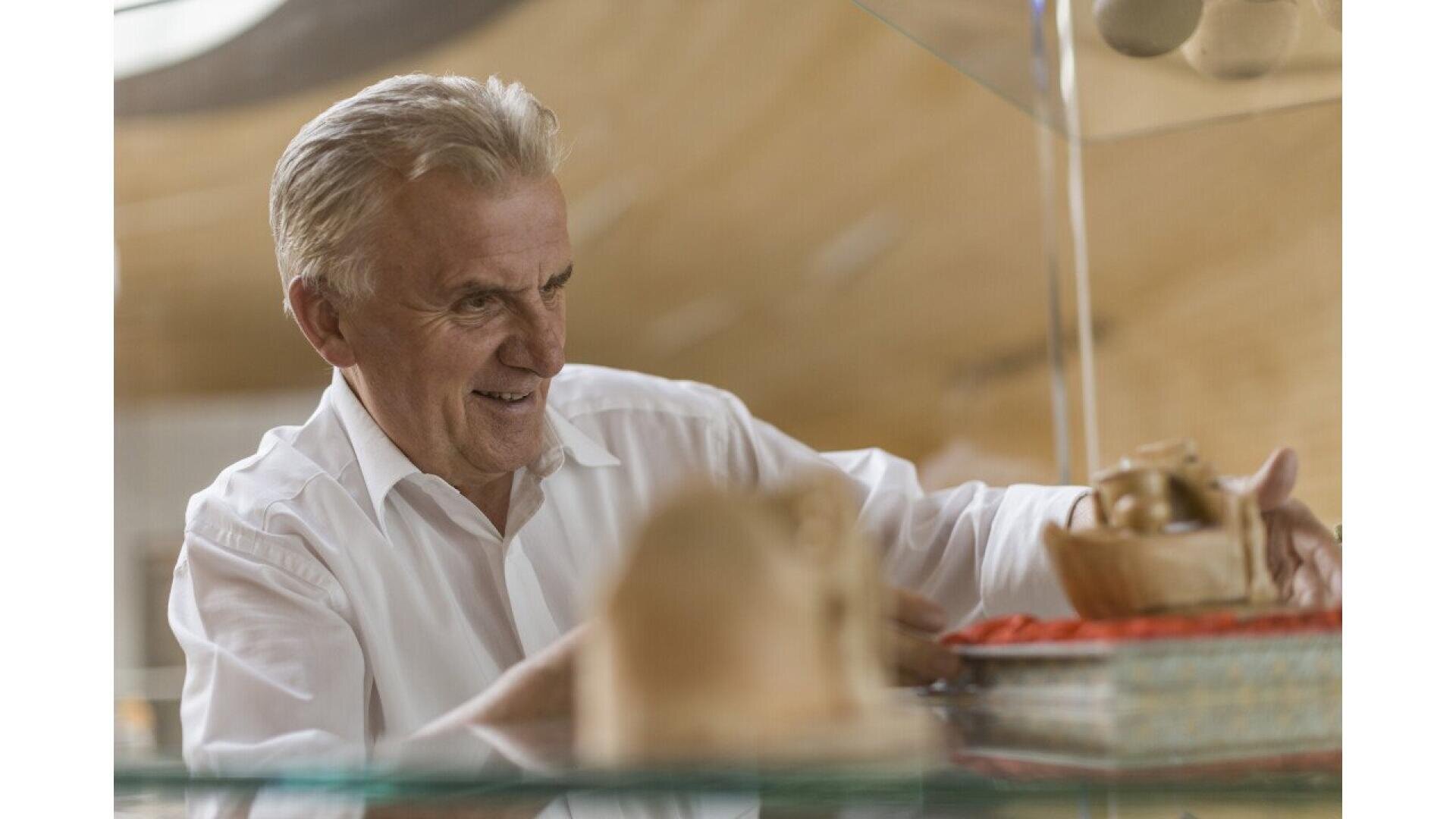 An older man with gray hair and a white shirt smiles while arranging wooden objects on a glass shelf in a warmly lit room.