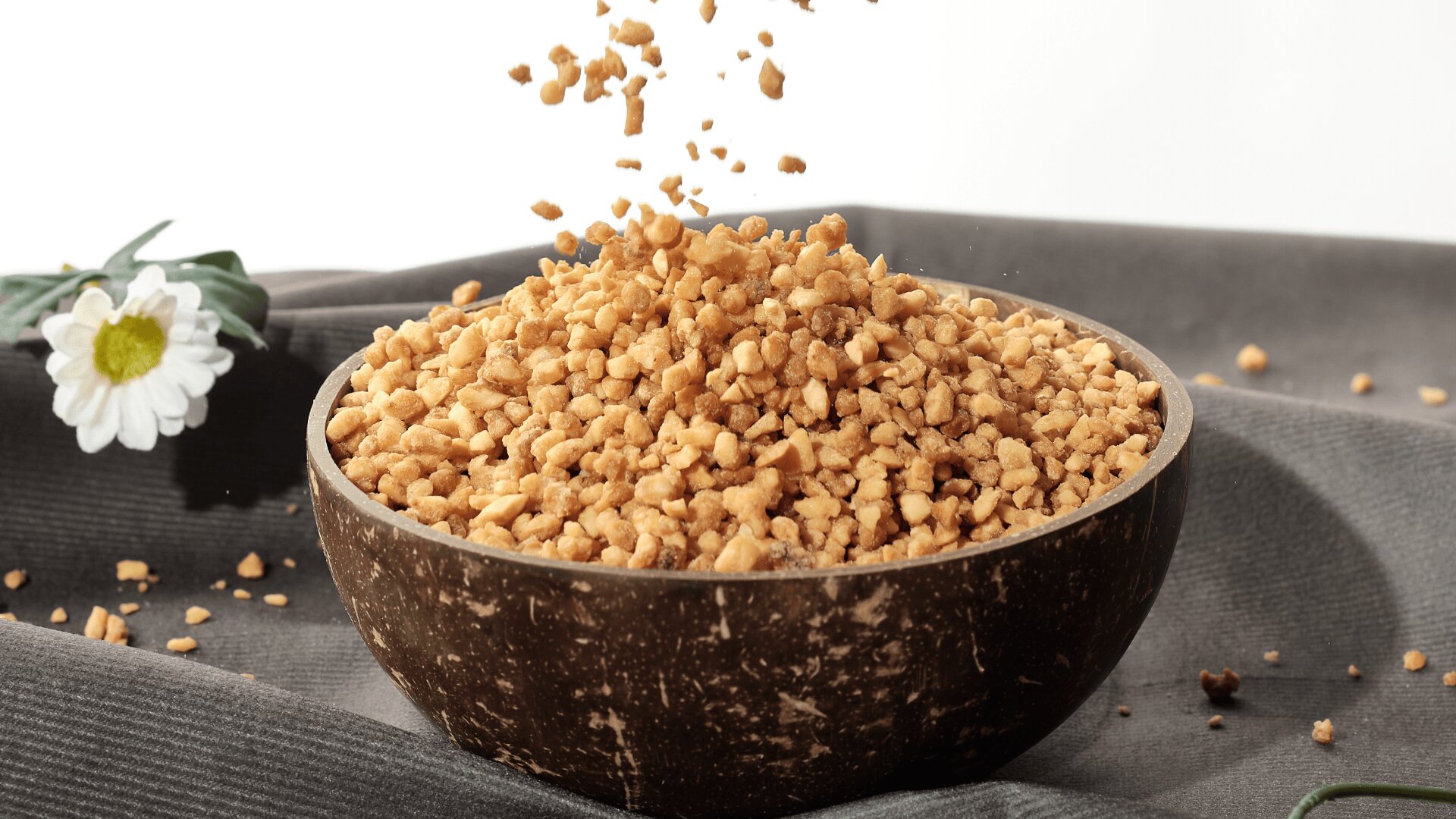 A wooden bowl filled with chopped nuts, with some pieces falling from above, sits on a gray fabric surface next to a white daisy flower.