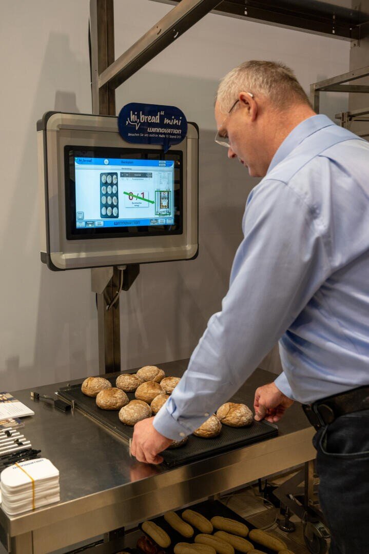 Ein Mann in einem blauen Hemd stellt ein Tablett mit runden Brötchen auf einen Tisch vor einem Touchscreen-Bedienfeld, das eine brotbezogene Schnittstelle in einer Großküche anzeigt.