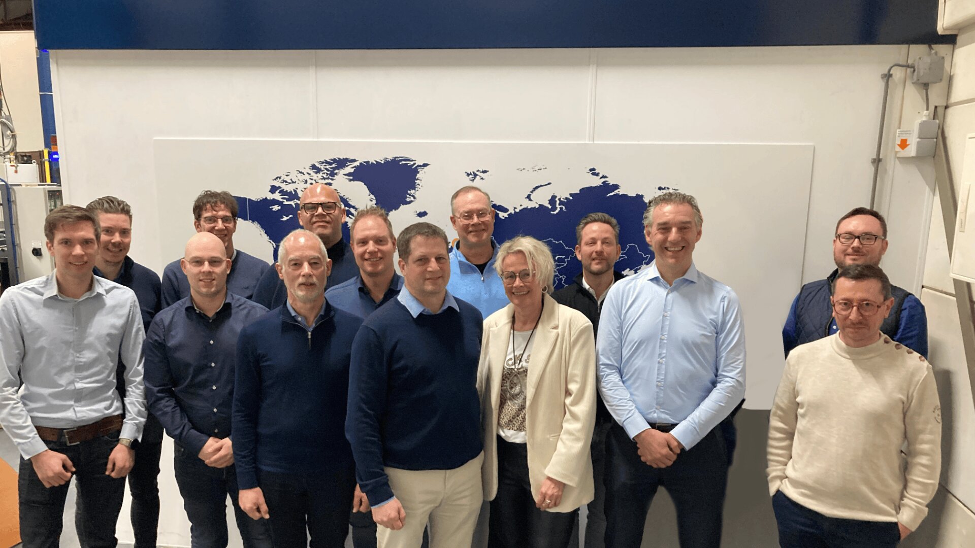 Eine Gruppe von dreizehn Personen, hauptsächlich Männer in Geschäftskleidung, steht lächelnd vor einem Wandgemälde mit einer Weltkarte an einer weißen Wand im Innenbereich.