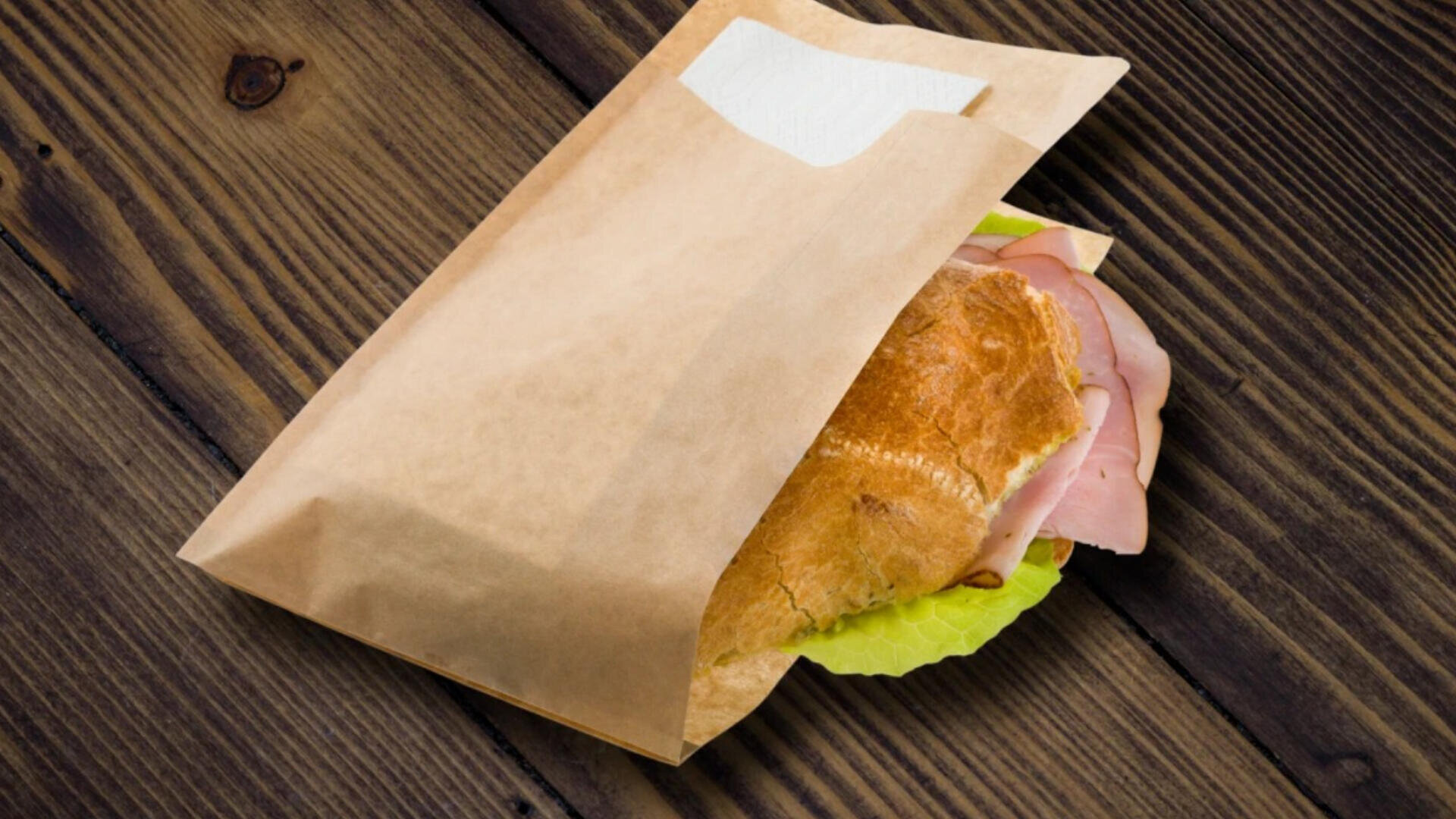 A sandwich with ham and lettuce is partially wrapped in a brown paper bag with a napkin, placed on a rustic wooden surface.