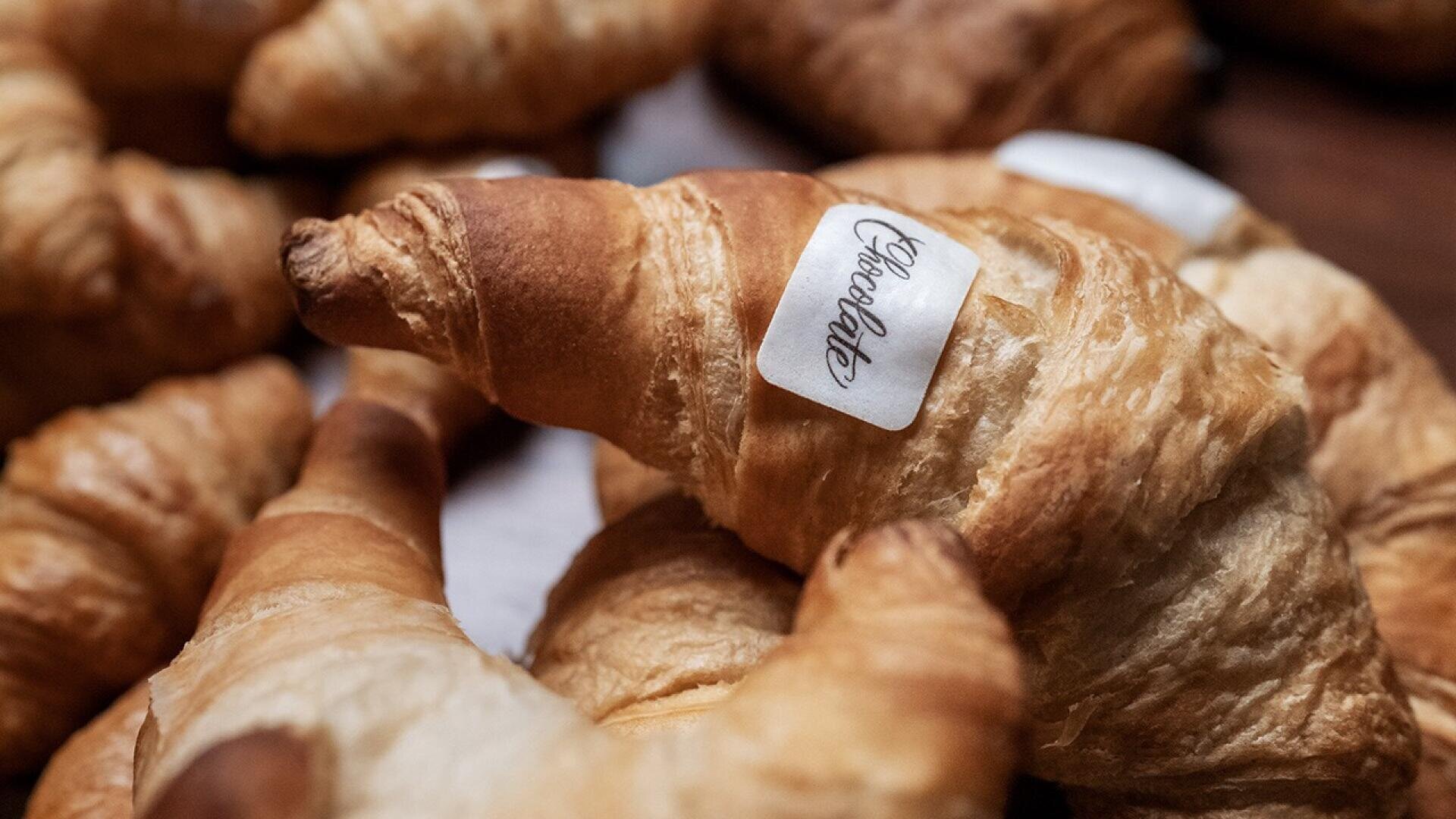 A close-up of several golden-brown croissants, with one in the foreground bearing a small white label that reads Croissant. The croissants are arranged on a wooden surface.