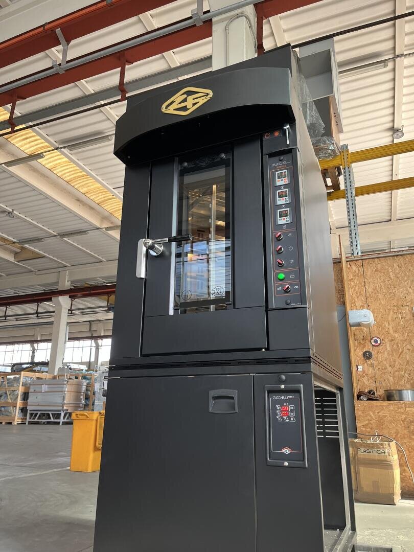 A large industrial black oven with a glass door and digital control panels stands in a spacious warehouse with high ceilings, exposed beams, and scattered equipment in the background.