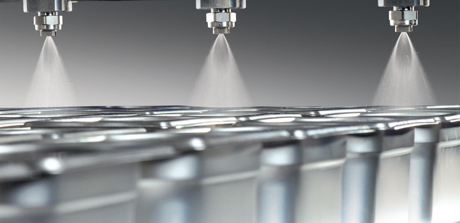 Nahaufnahme von Sprühdüsen, die einen feinen Nebel über Reihen von Metalldosen oder -behältern abgeben, wahrscheinlich Teil eines industriellen Herstellungs- oder Beschichtungsprozesses.