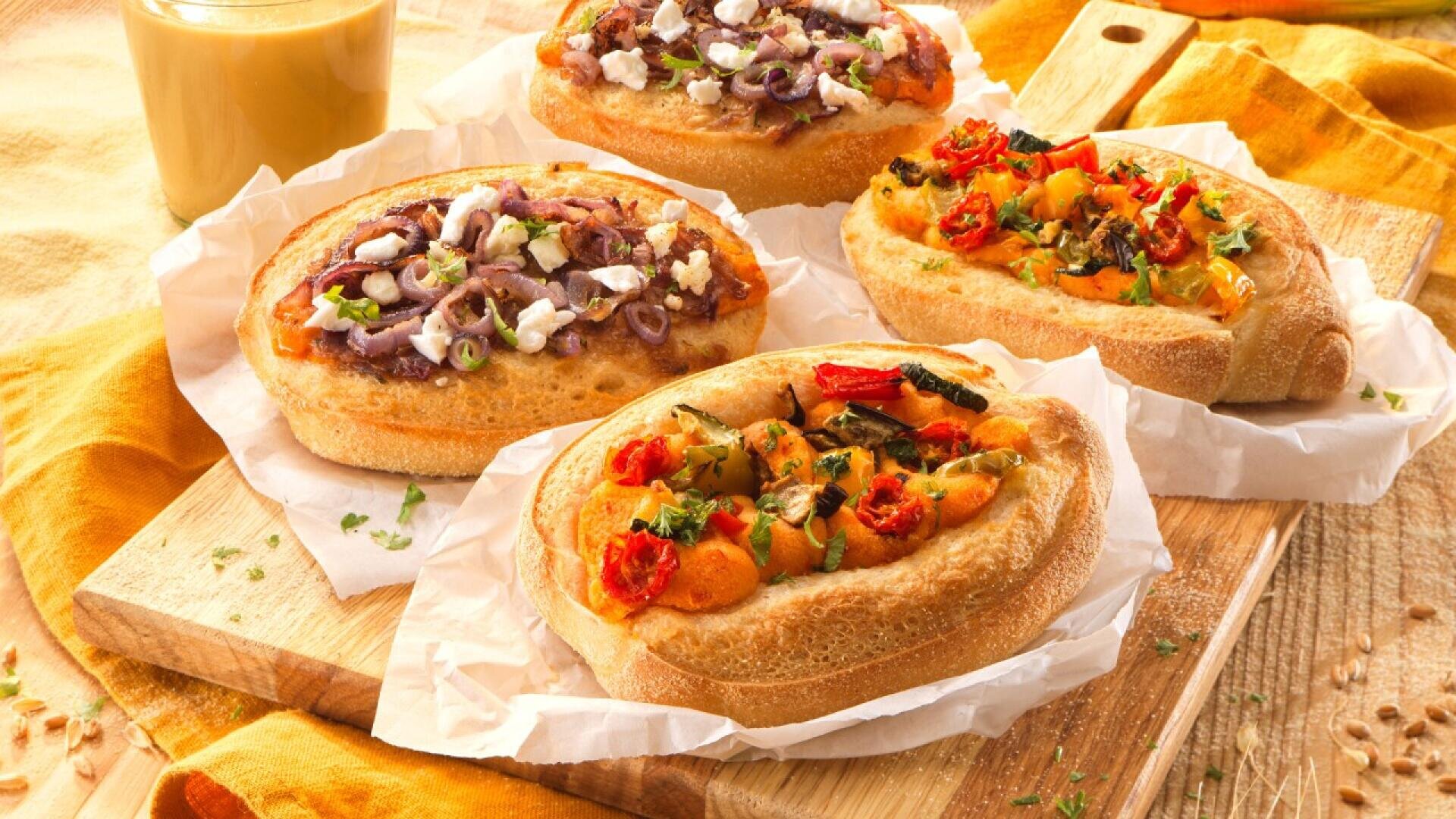 Four oval-shaped mini breads topped with colorful vegetables, cheese, and herbs are placed on parchment paper over a wooden board, with a glass of orange juice in the background.