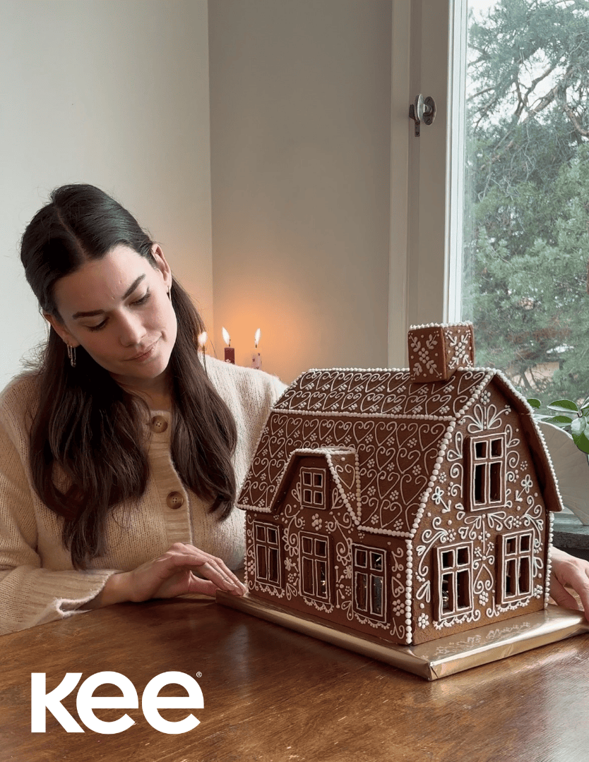 A woman in a beige sweater admires a large decorated gingerbread house on a wooden table. Lit candles and a window with trees outside are in the background. The word “kee” is in the bottom left corner.
