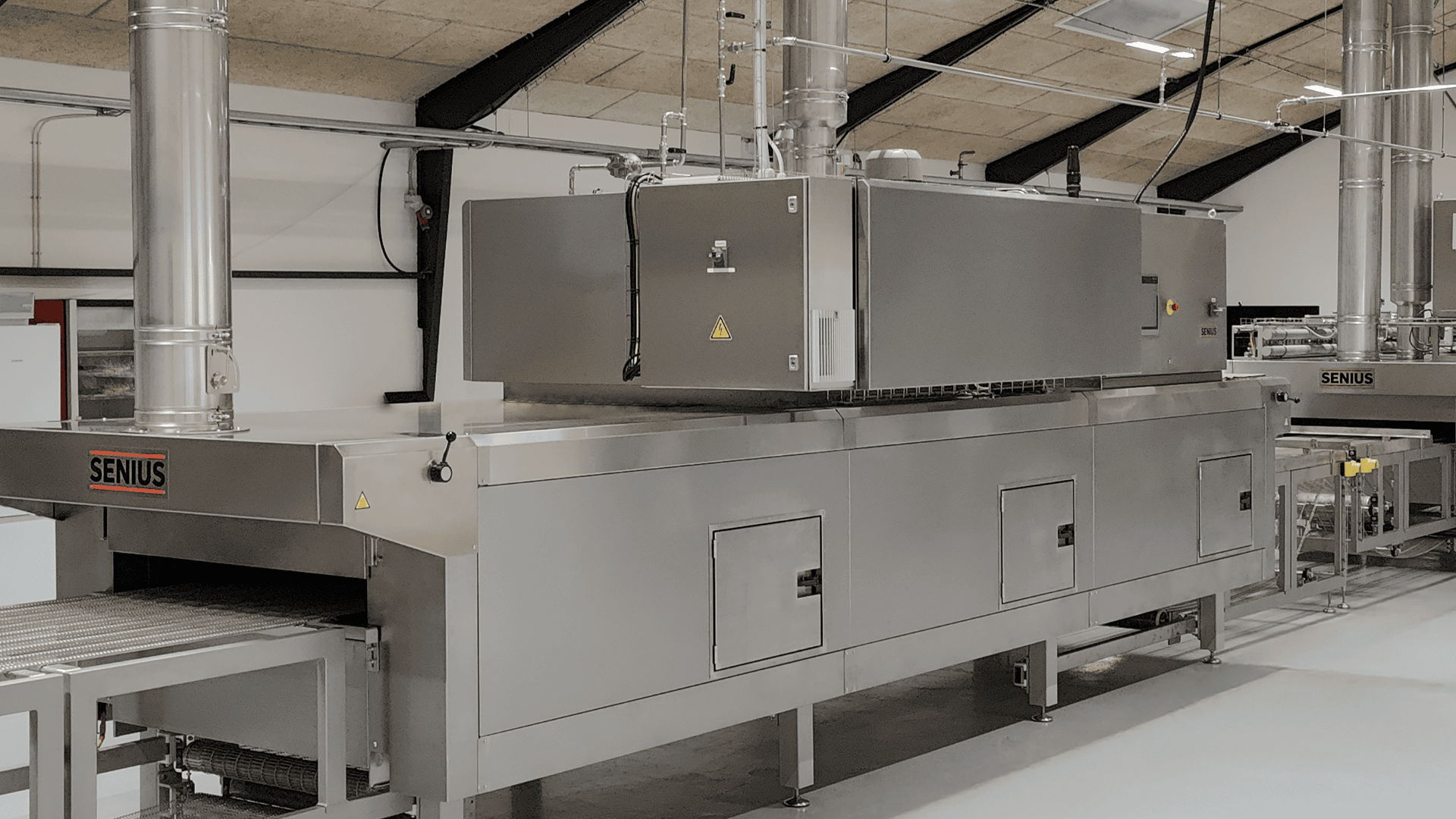 A large industrial stainless steel oven with conveyor belts, vents, and control panels in a modern factory setting. The brand SENIUS is visible on the machines side.