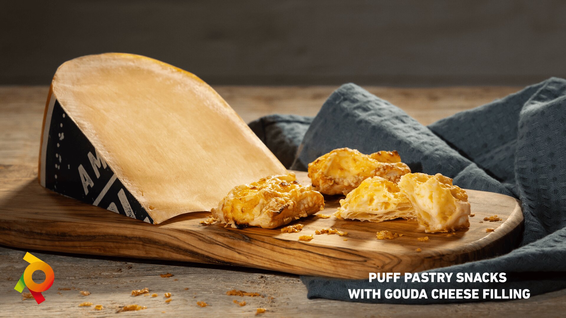 A wedge of Gouda cheese and three golden puff pastry snacks are arranged on a wooden board beside a blue cloth. Crumbs are scattered on the board. Text reads: PUFF PASTRY SNACKS WITH GOUDA CHEESE FILLING.