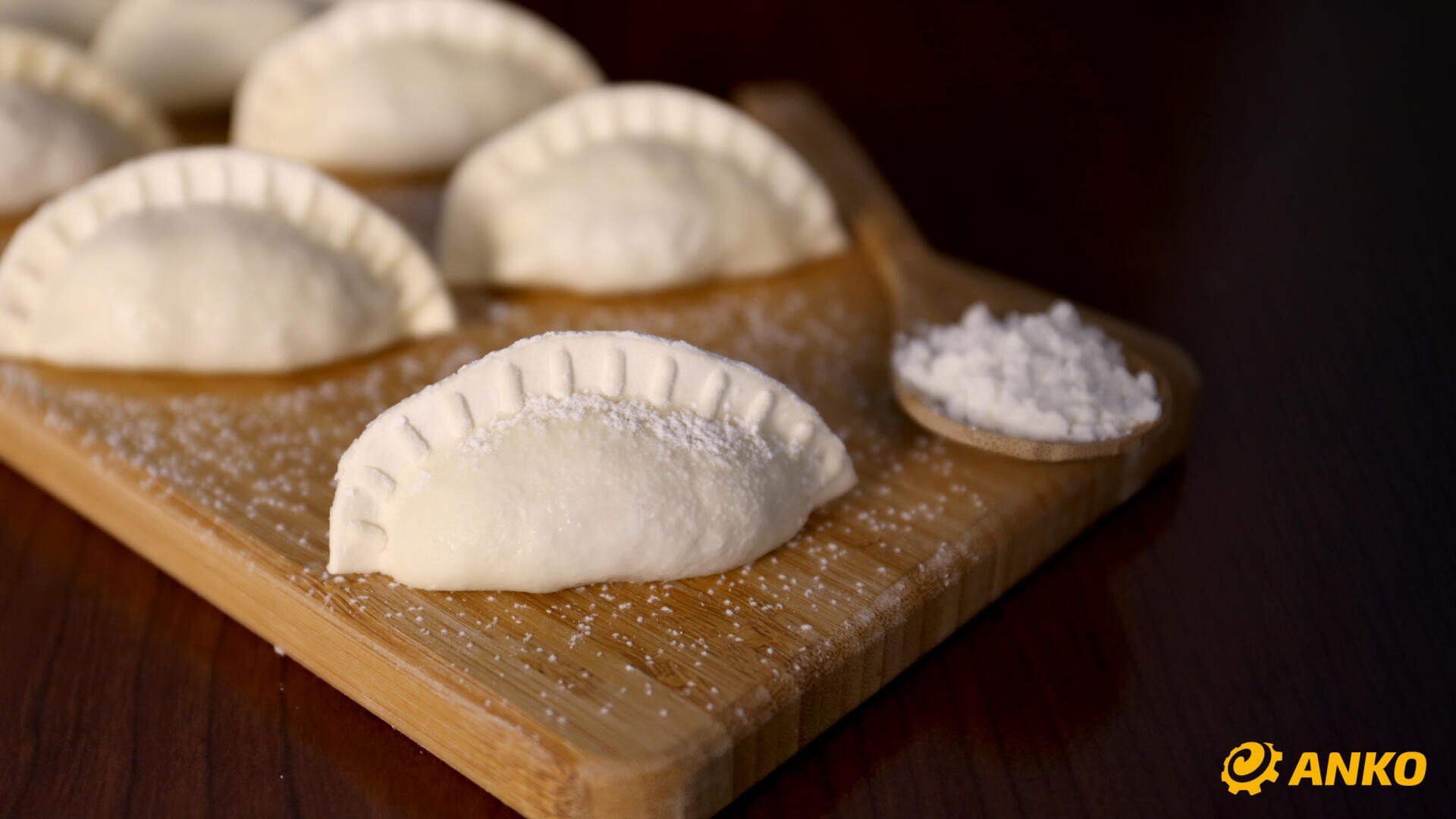 Six uncooked dumplings are arranged on a wooden cutting board, with a small wooden spoon filled with flour beside them. The ANKO logo is visible in the bottom right corner.
