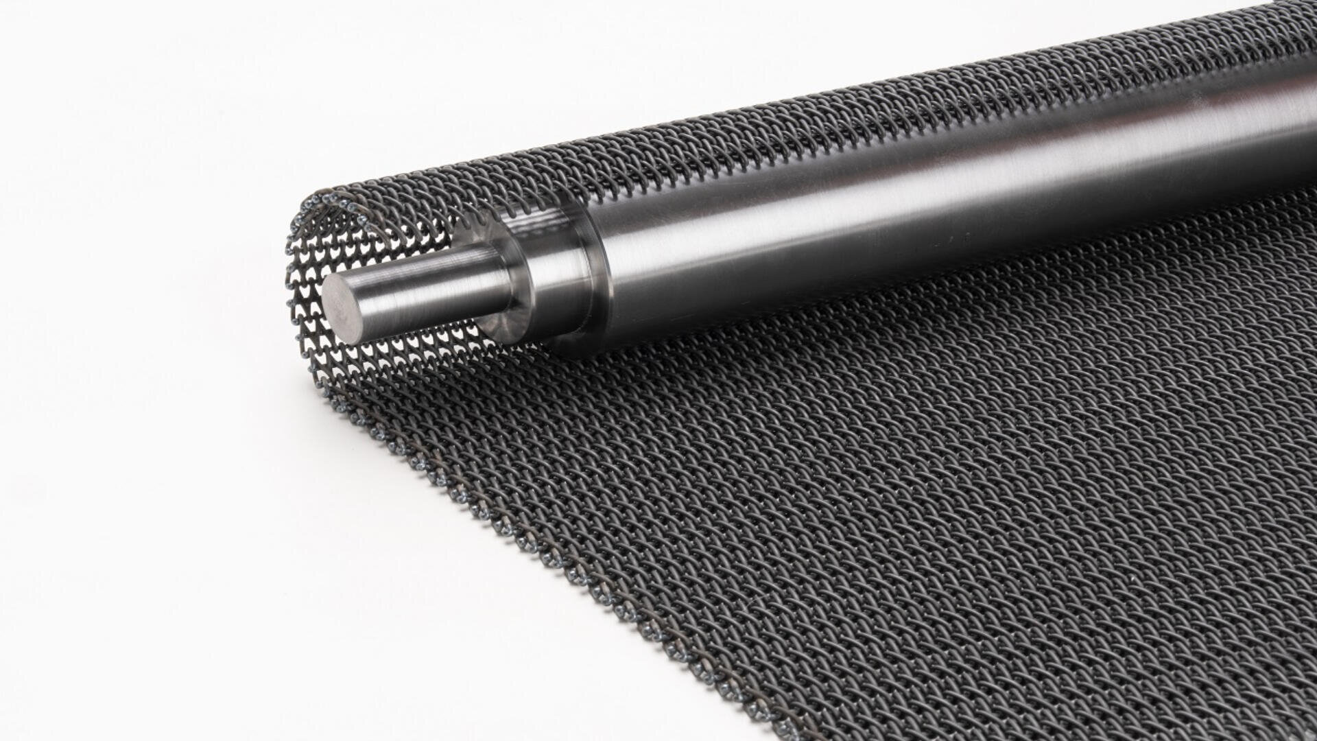 A close-up of a black metal mesh conveyor belt rolled around a cylindrical rod on a white background.