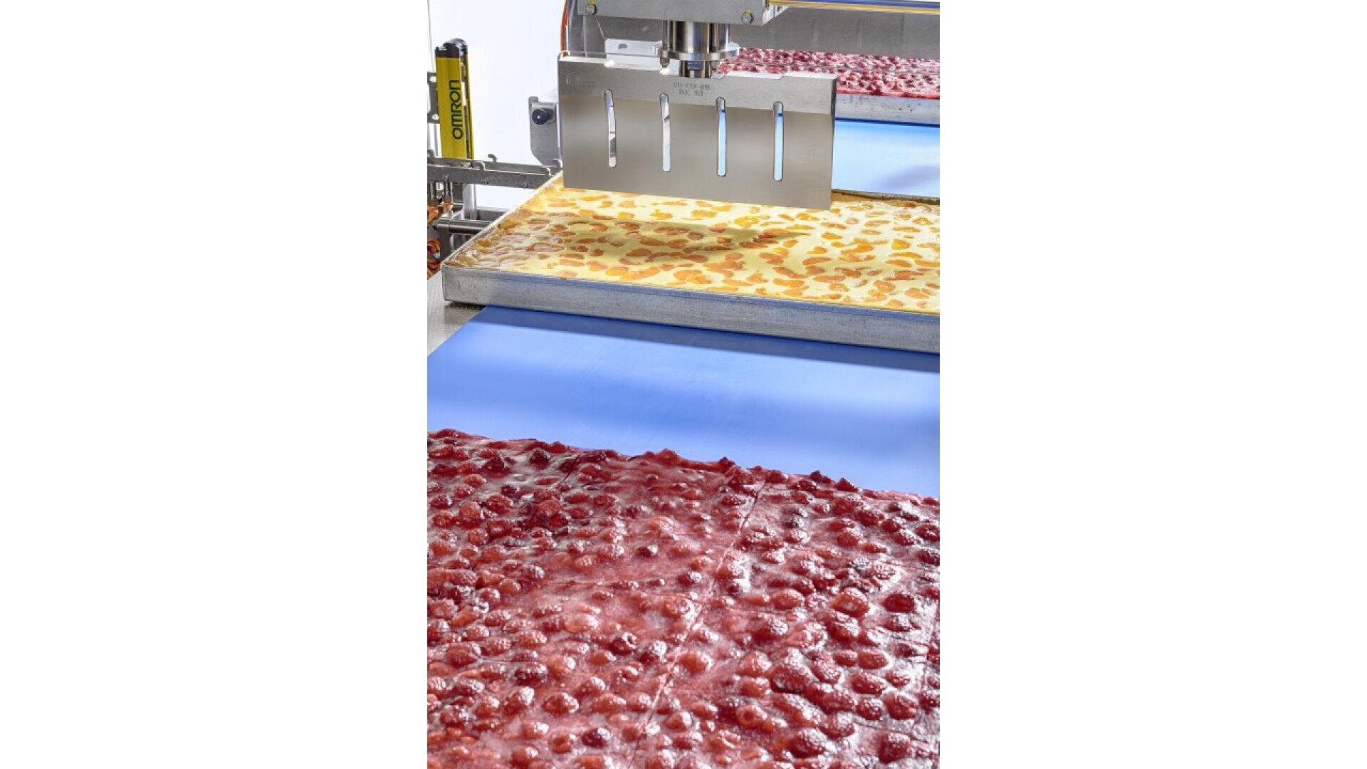 A food processing machine slices large sheets of pizza or pastry dough topped with cheese and diced vegetables on a blue conveyor belt. Another sheet with red sauce and toppings is in the foreground.