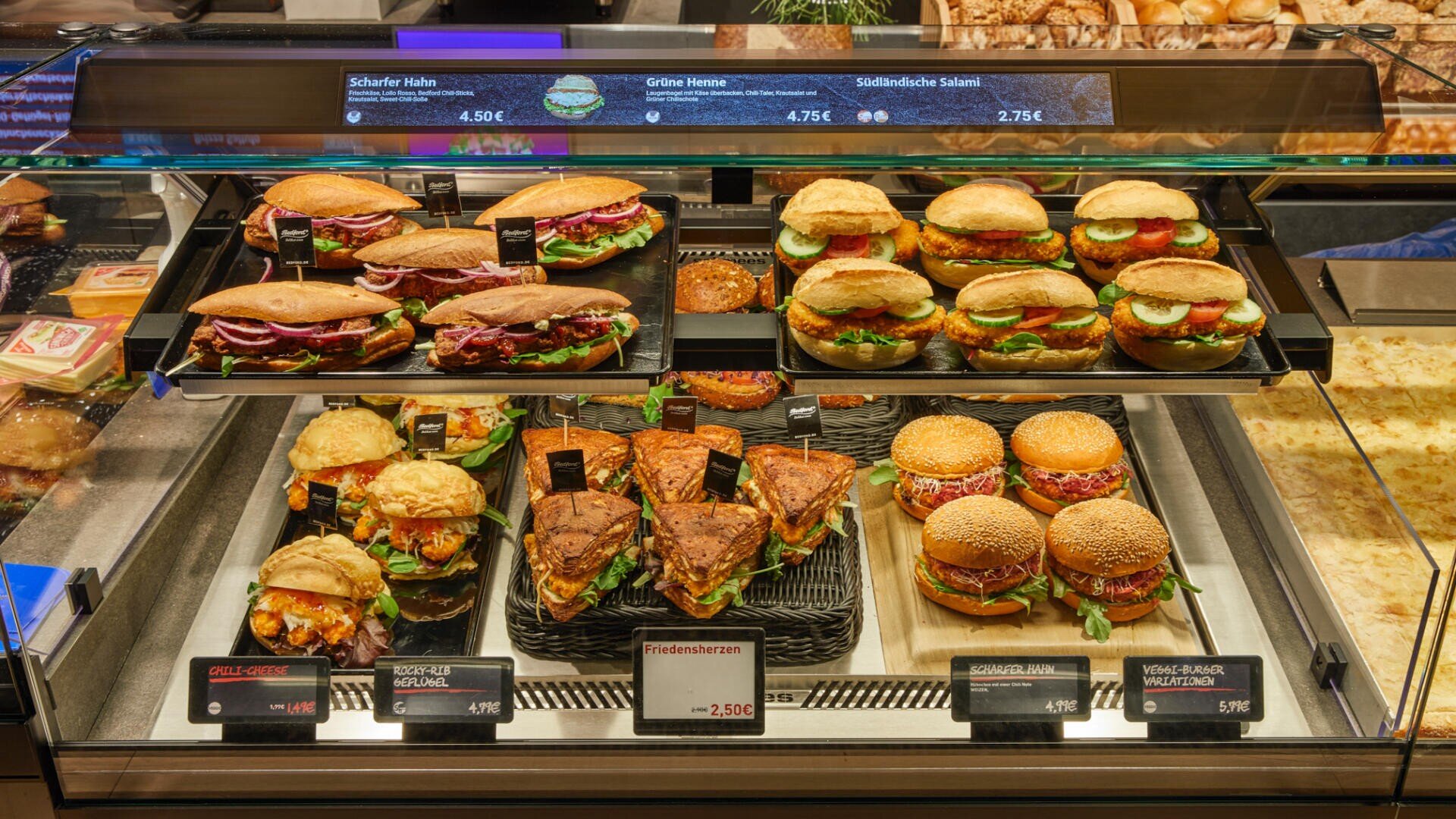 Eine Bäckereivitrine, gefüllt mit verschiedenen Sandwiches und Burgern auf verschiedenen Brotsorten, die jeweils mit Namens- und Preisschildern versehen sind. Der Hintergrund zeigt eine Vielzahl von Backwaren wie Brot und Brötchen.