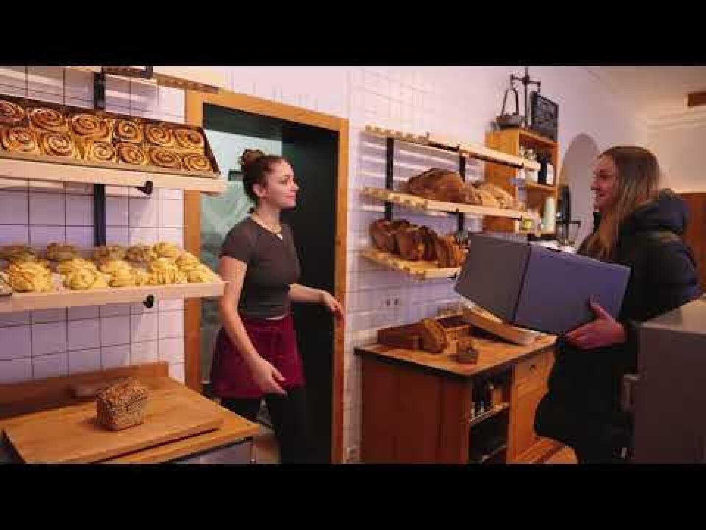 A woman behind the counter at a bakery talks to a customer holding a large box. Shelves filled with bread and pastries are visible in the background.