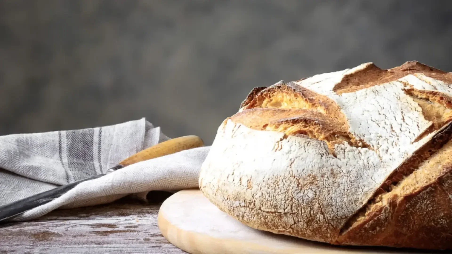 A round loaf of rustic sourdough bread sits on a wooden board. A cloth napkin and a serrated knife are placed to the left on a weathered wooden surface, with a soft, neutral background.